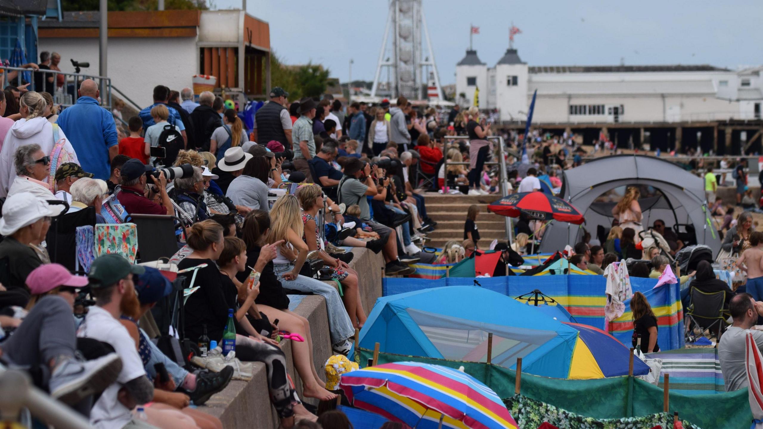 A large crowd of people watching the show on the seafront in Clacton. They are either standing above the concrete sea wall or sitting on camping chairs on the beach.