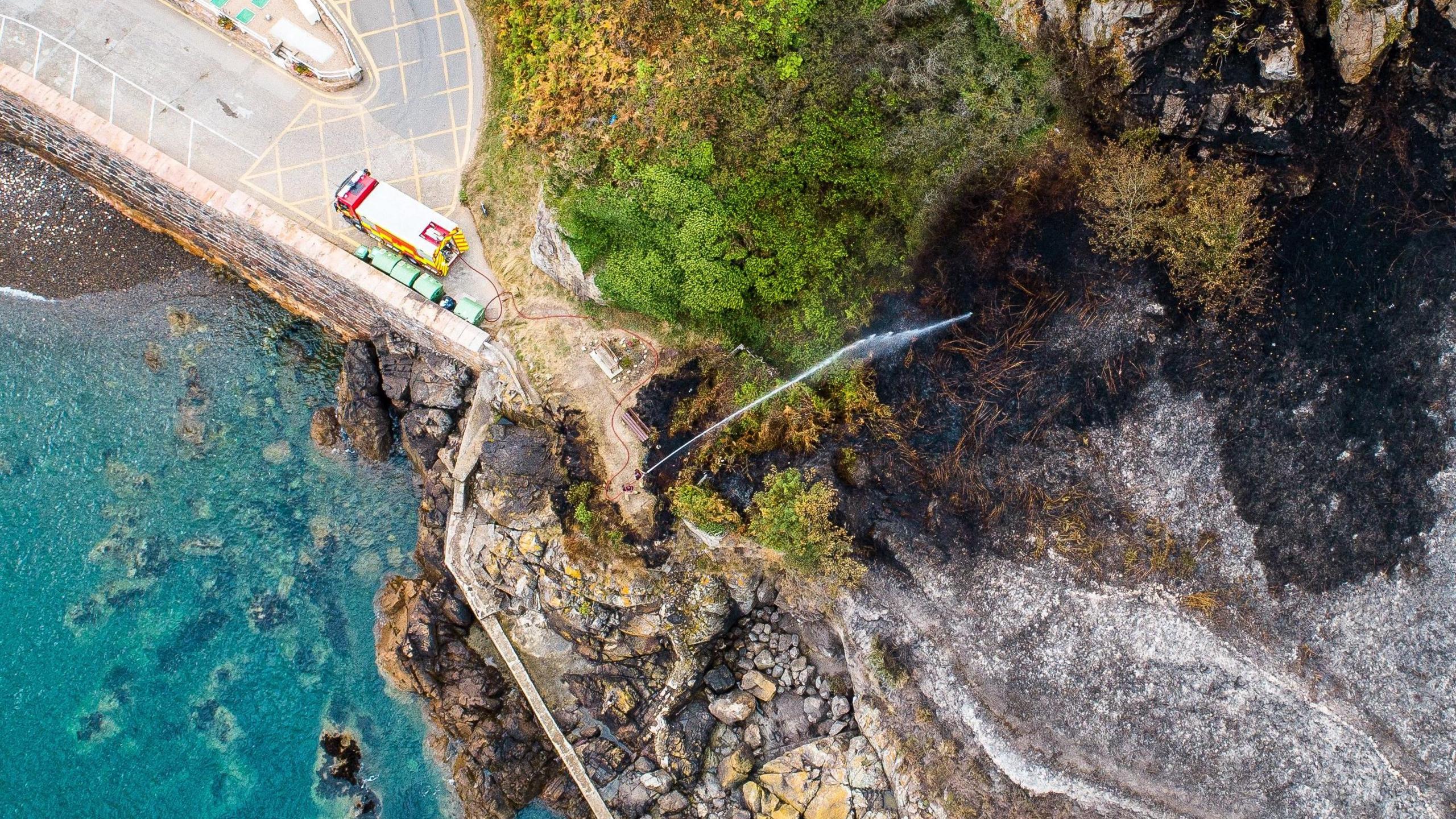 Aerial view of wildfire at Bonne Nuit in 2018. A fire engine is parked in a car park next to the open land which had been damaged by the flames. Firefighters are standing on the edge of the land, next to the water, using a hose reel jet to spray water over the area.
