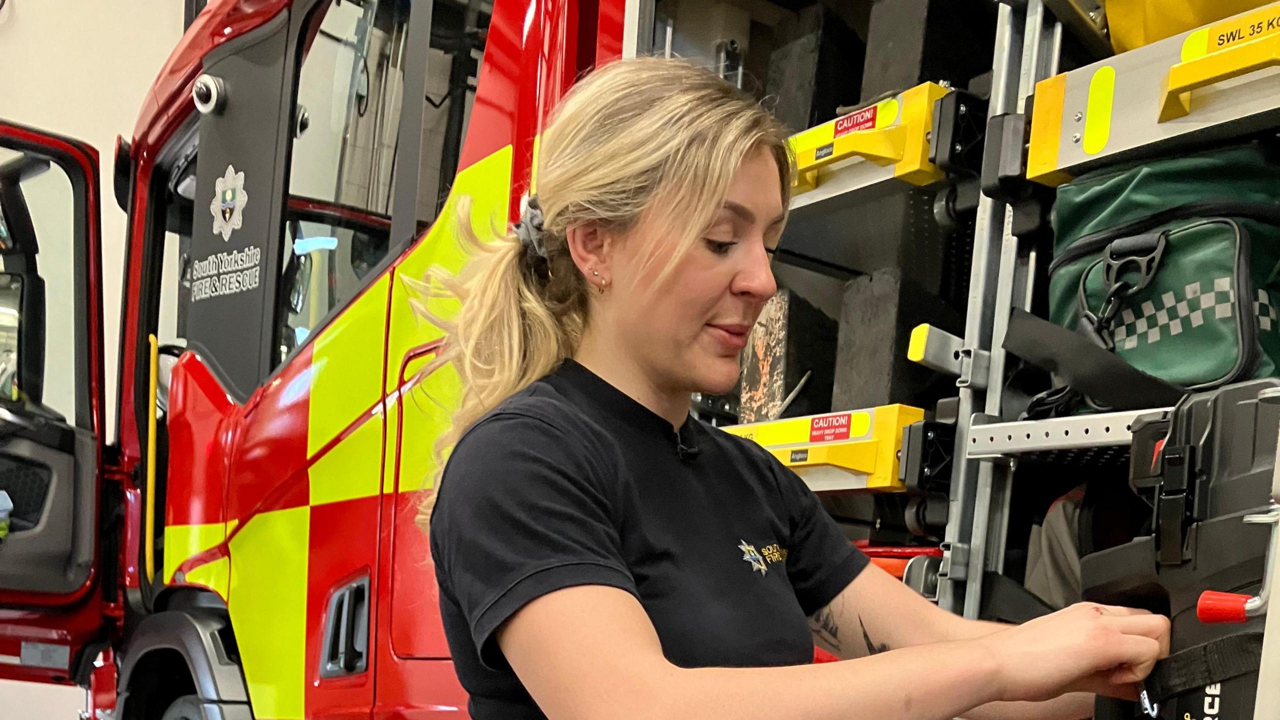 Bronte Jones, pictured against the backdrop of her fire station, which includes a fire engine and some shelves. She appears to be looking inside some kind of drawer.