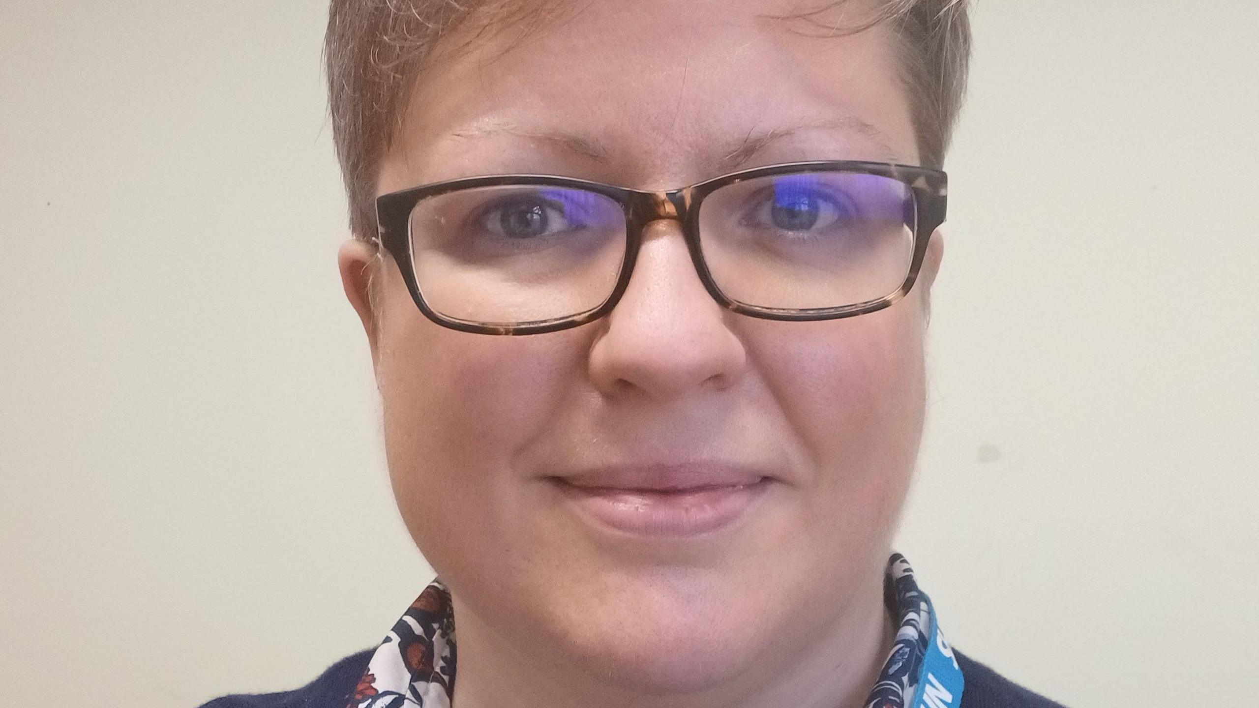 A woman with short blonde hair, glasses and wearing a shirt and jumper looks at the camera