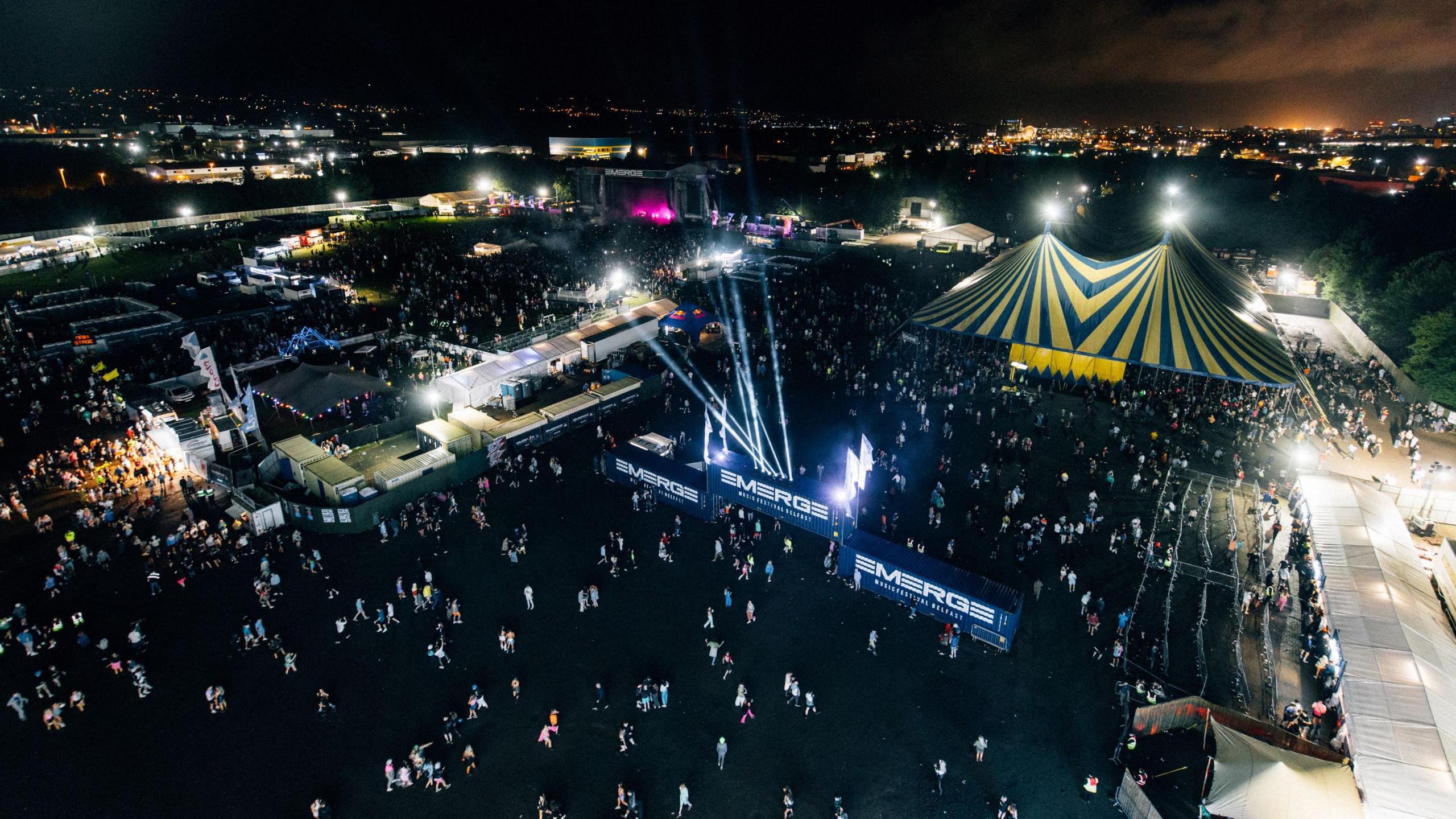 Aerial view of Emerge from last year. There are lots of people. The tents and main stage are lit up and you can see the Belfast skyline in the background.