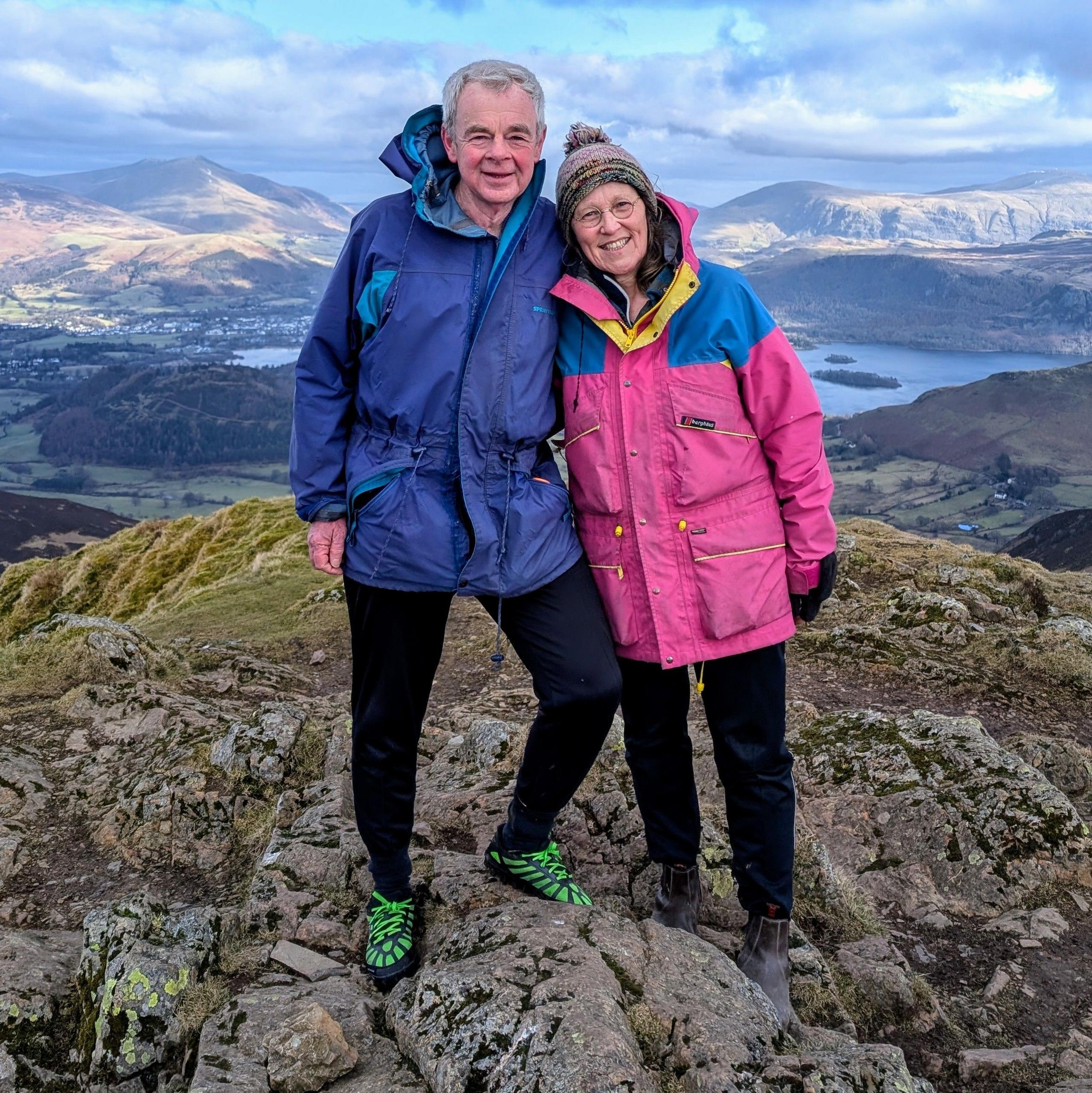 Ian Teasdale and Catherine Wetherfield standing on top of a hill. There are wearing outdoor coats. Ian's is blue and Catherine's is pink. She also wears a bobble hat. They are both smiling. In the distance behind them is a lake and more mountains. The ground beneath them is rocky. The both wear specialist walking shoes. Ian's are green.