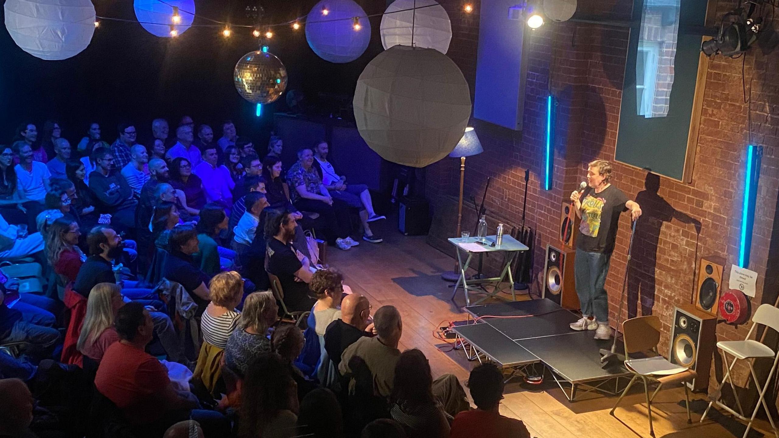Josie Long, dressed in jeans and a black T-shirt, holding a microphone while stood on stage in front of a crowd.