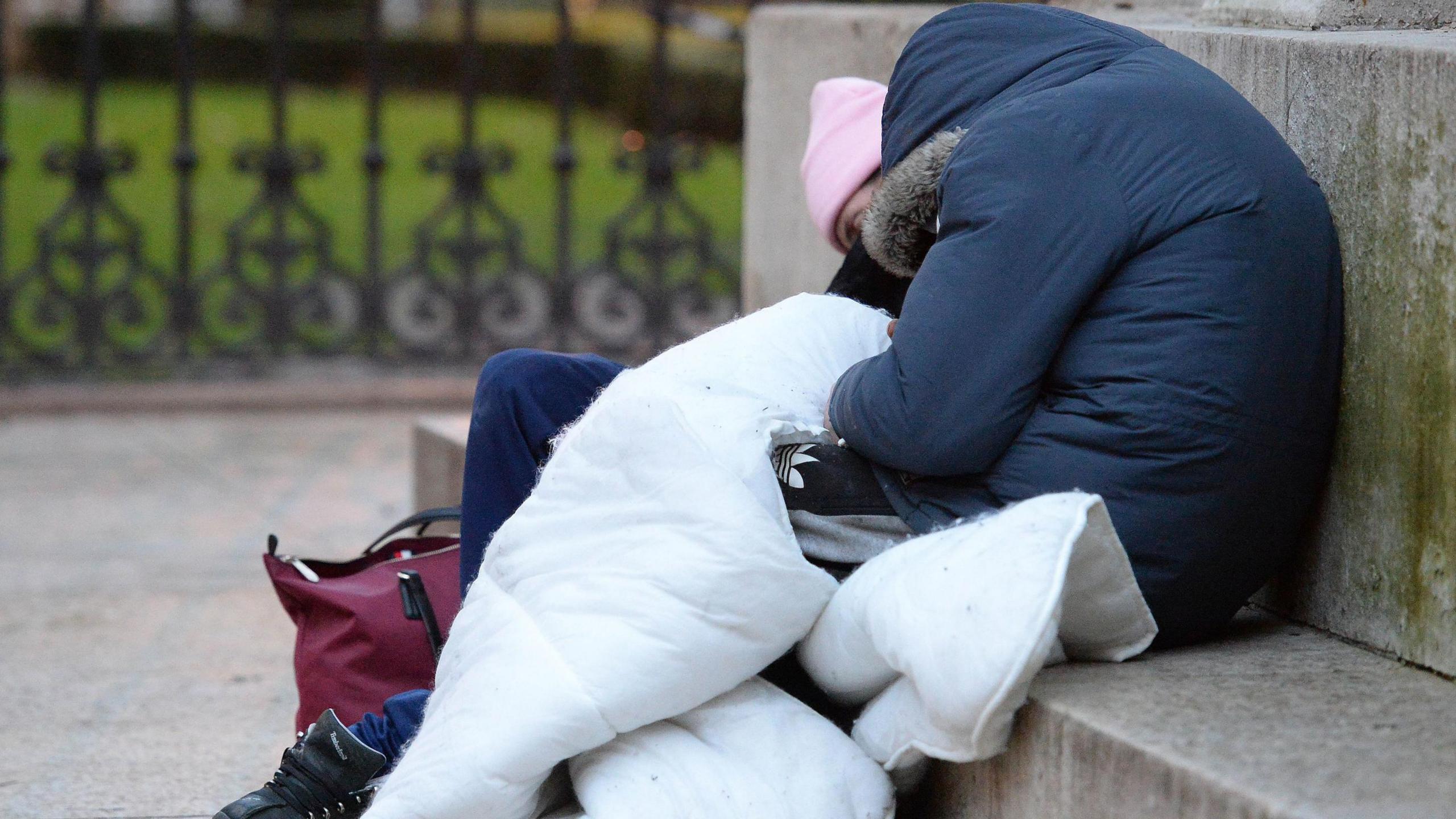 Two figures wrapped in thick coats and blankets sleep on a concrete step. 