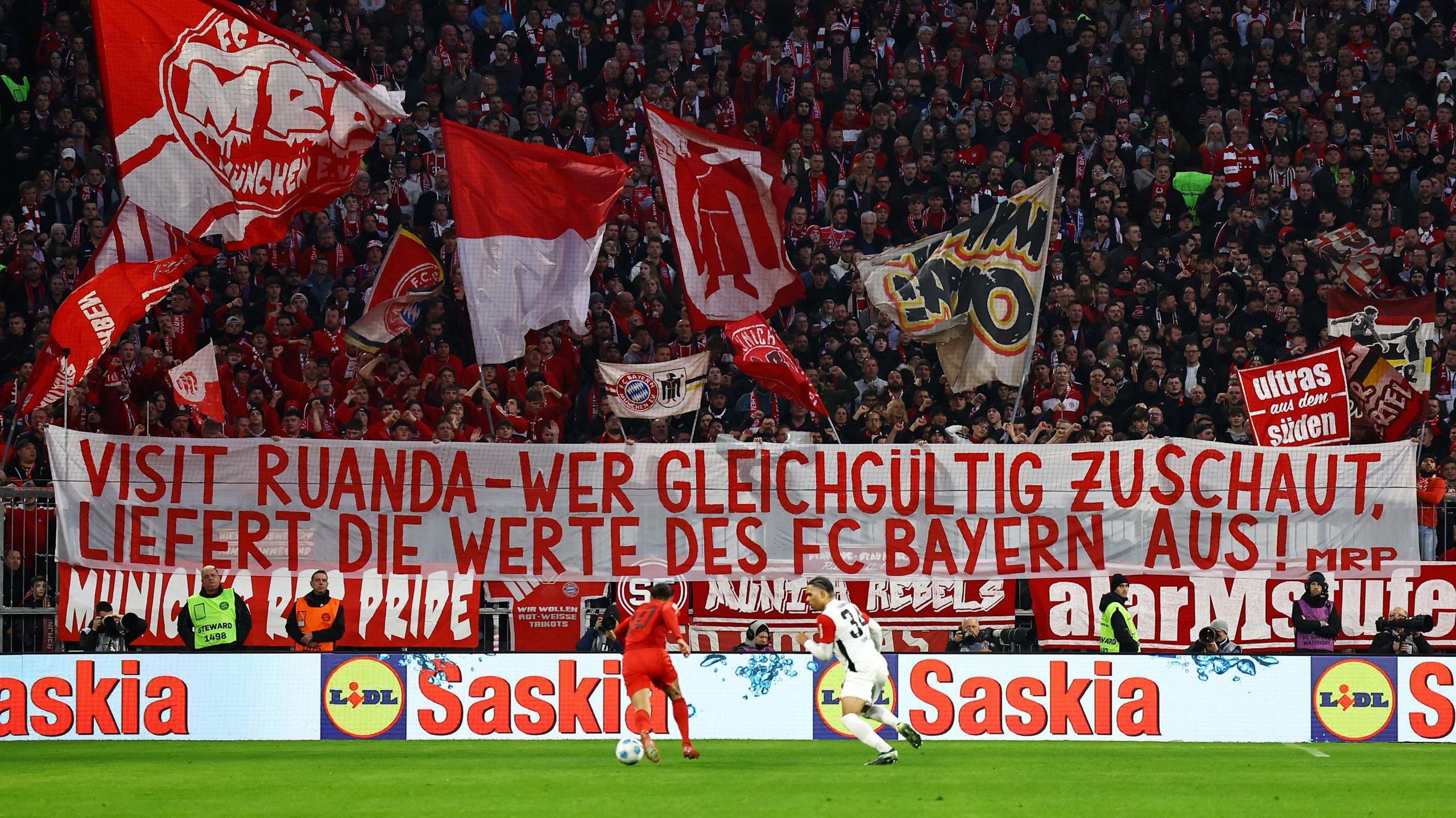 Bayern Munich fans hold up a banner in German which translates as 'Visit Rwanda - Whoever looks on with indifference is betraying the values of FC Bayern' during a football match as two players are seen on the pitch in the foreground