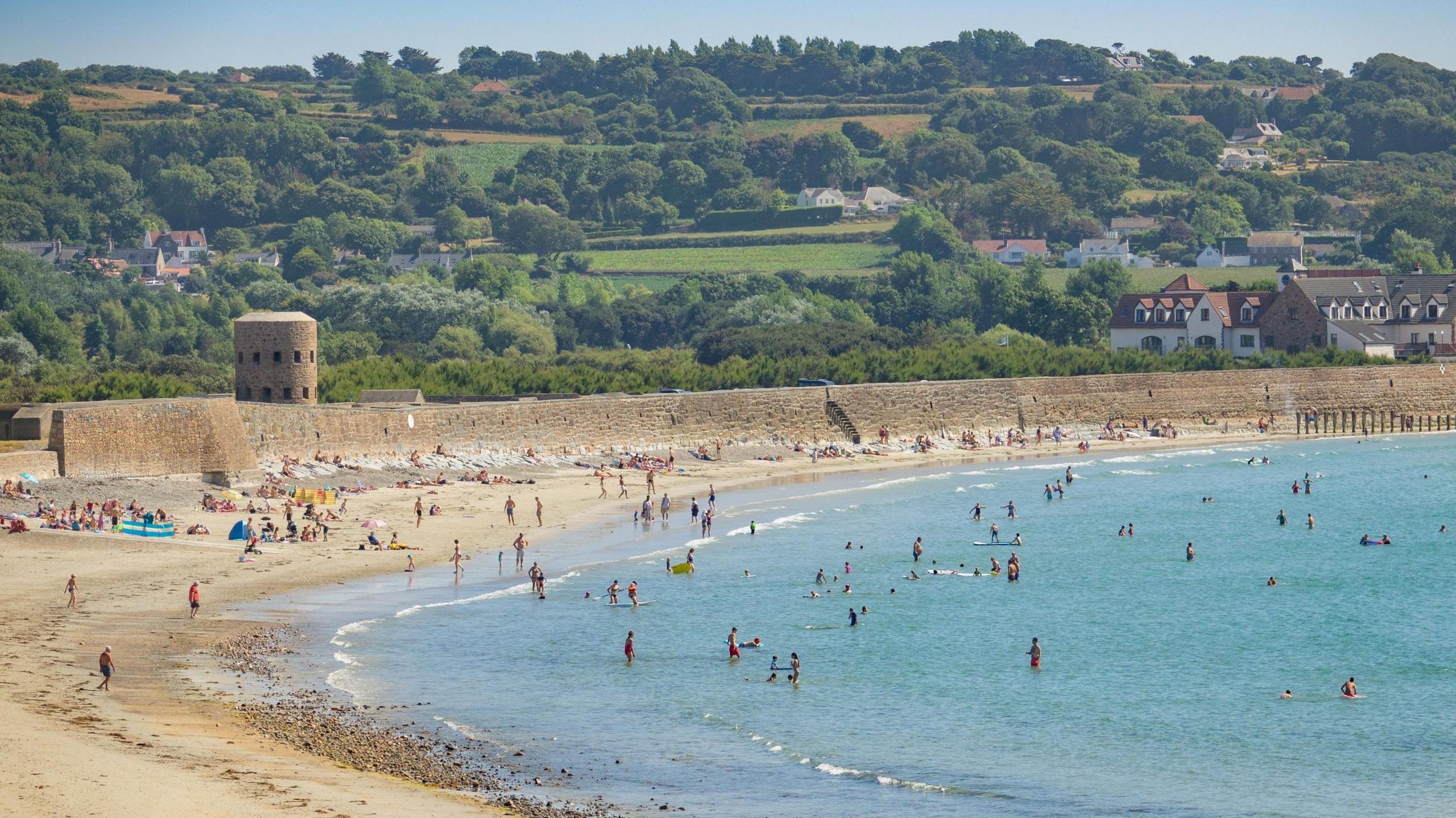 The beach and sea at Vazon Bay is full of beachgoers on a sunny day with green fields, trees and a few houses on the hills in the background.