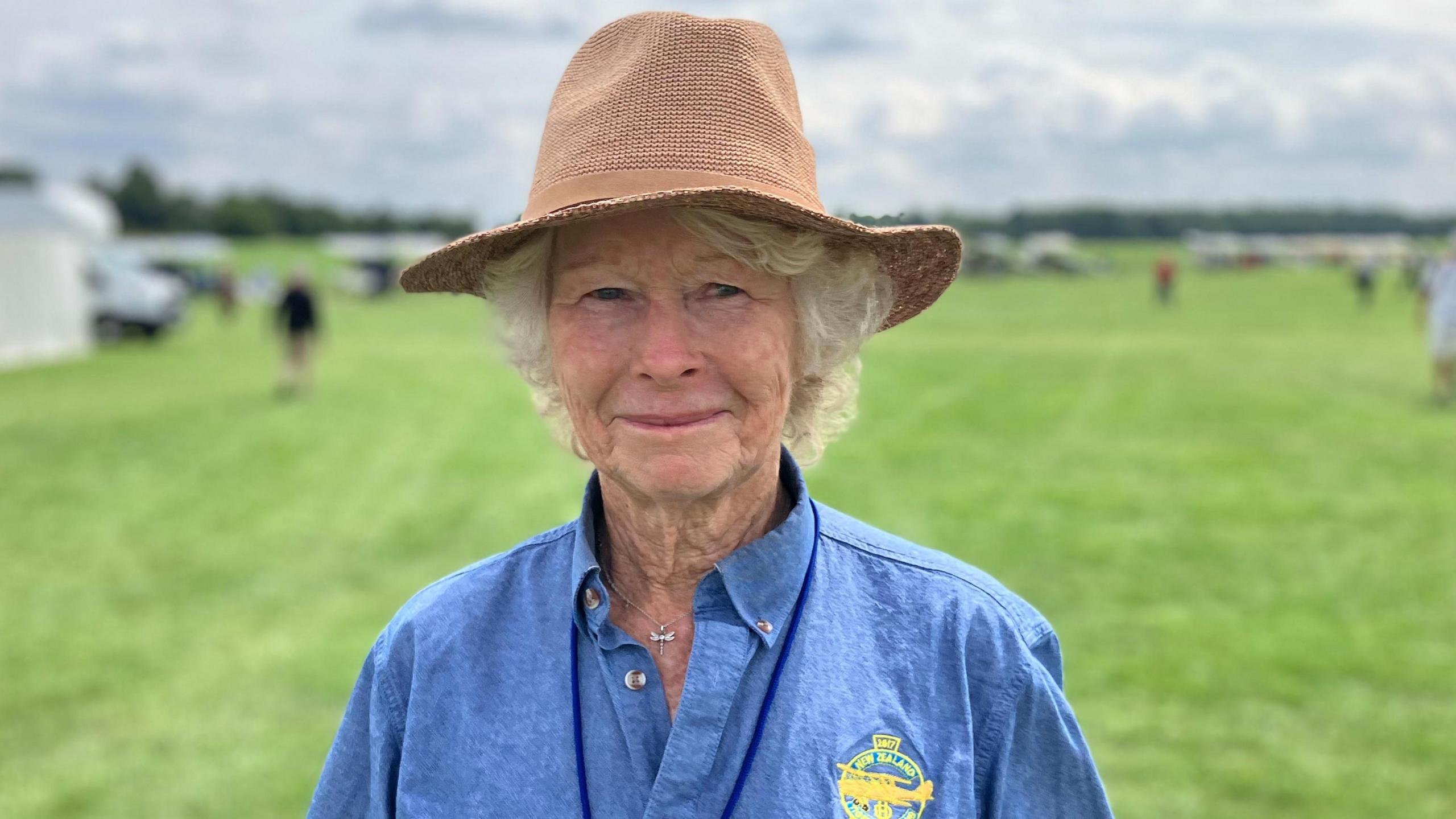 An older woman standing on a grassy field wearing a brown hat and a blue shirt. Other people are in the background, under a cloudy sky.