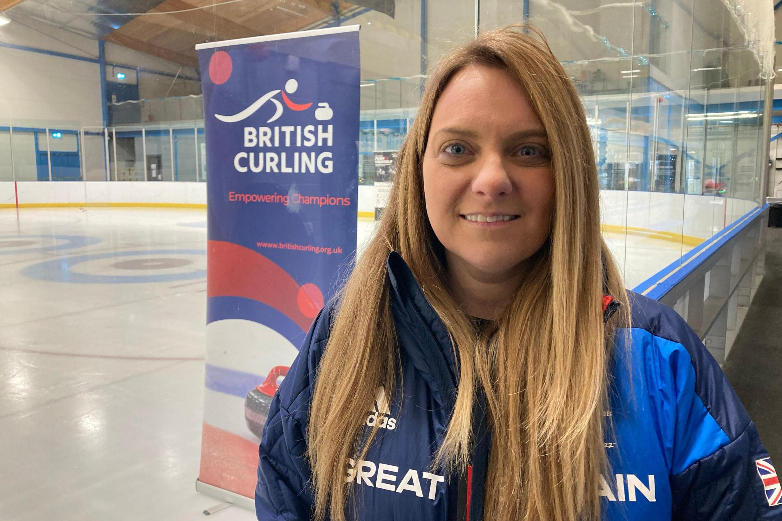 Cheryl Lappin from British Curling is looking at the camera, wearing a blue jacket with Great Britain written across the front. She has long, fair hair and is standing at the side of an ice rink with a British Curling banner behind her