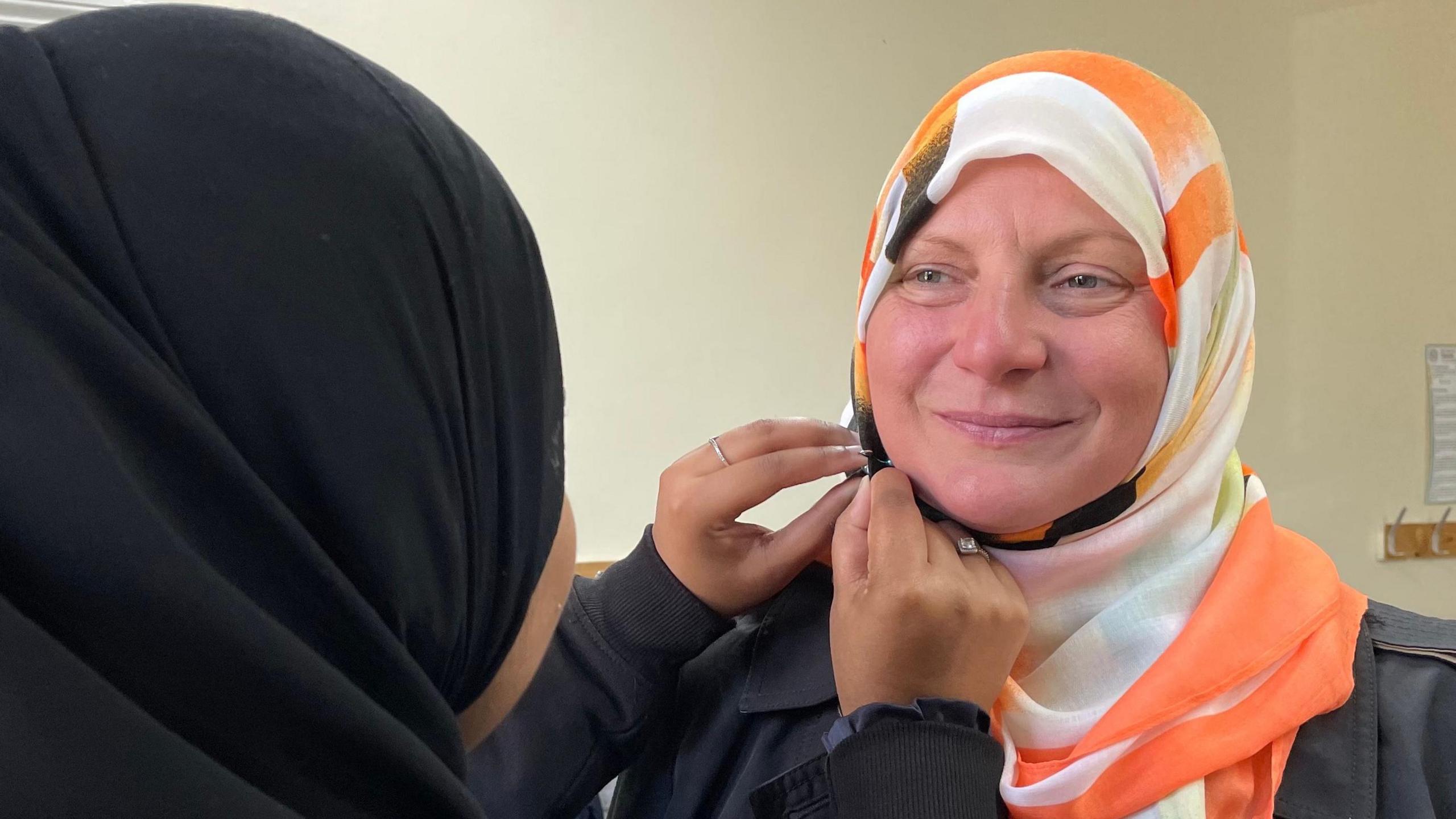 A woman smiles as another woman, who is wearing a black headscarf, helps her tie an orange and white hijab.