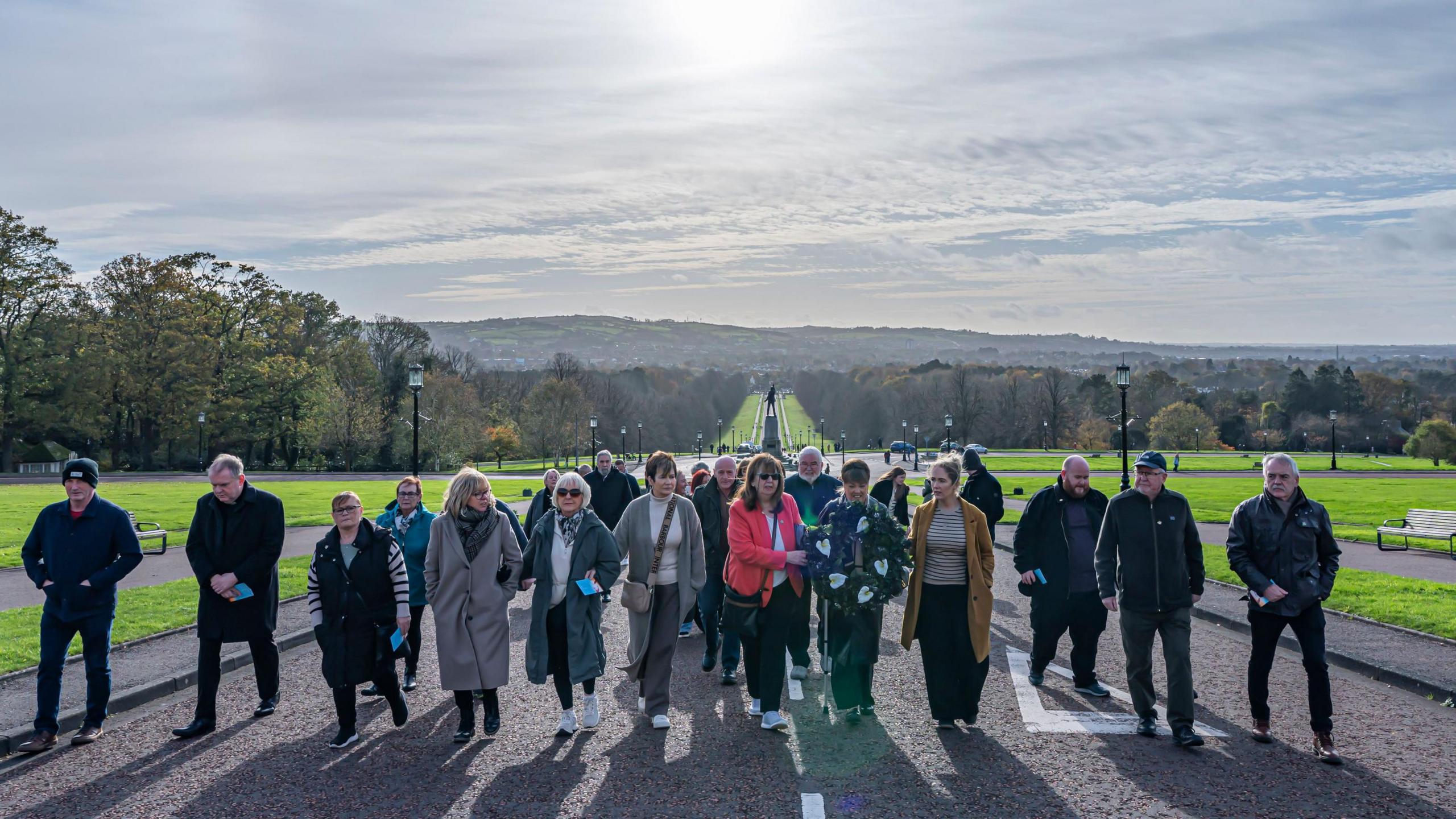 Families of the Disppeared walk through the grounds of Stormont on a sunny day carrying a large black wreath.  A lawned avenue stretches down the hill behind them. 