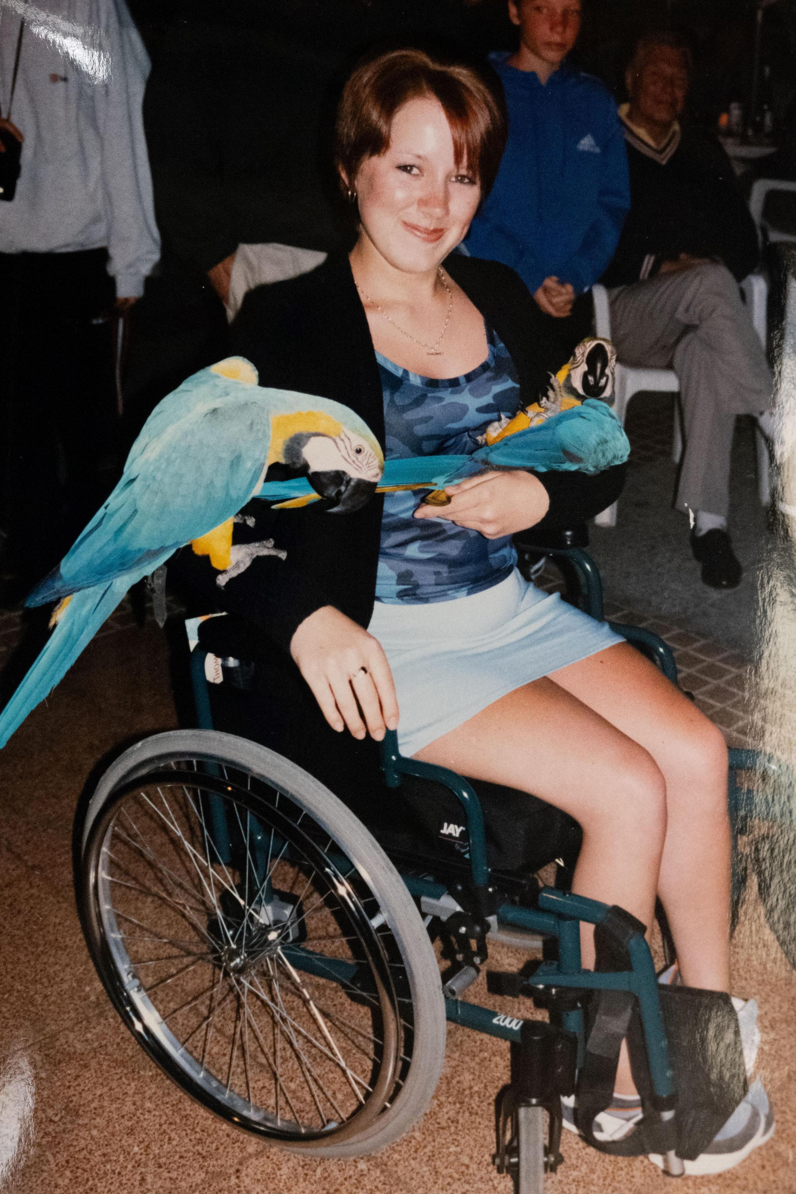 Amy Stevenson in her teenage years. She has short brown hair and sits in a wheelchair with two parrots perched on her arms.