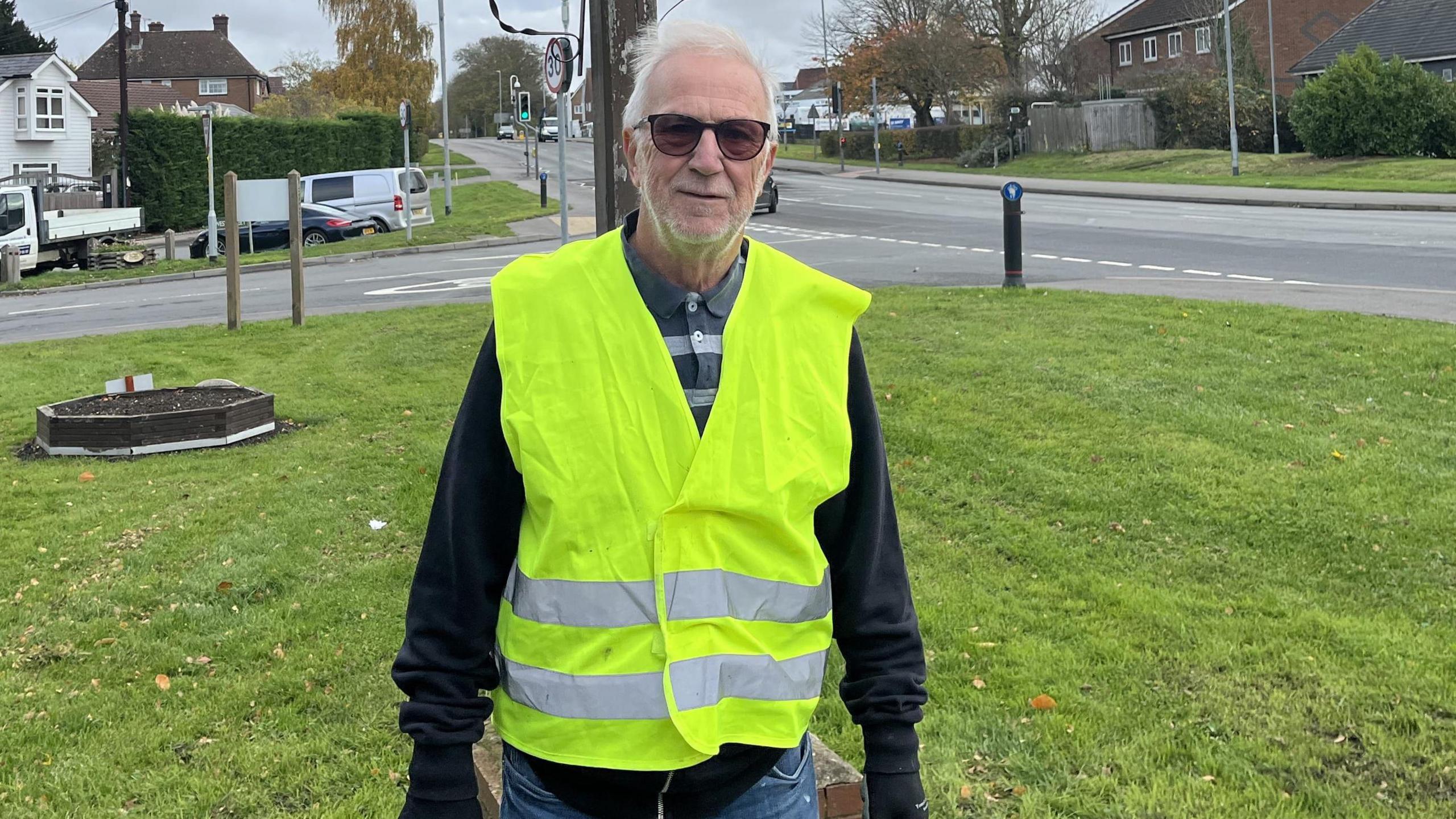 A man in a yellow high vis jacket standing on a patch of grass. He has short white hair and is wearing sun glasses.