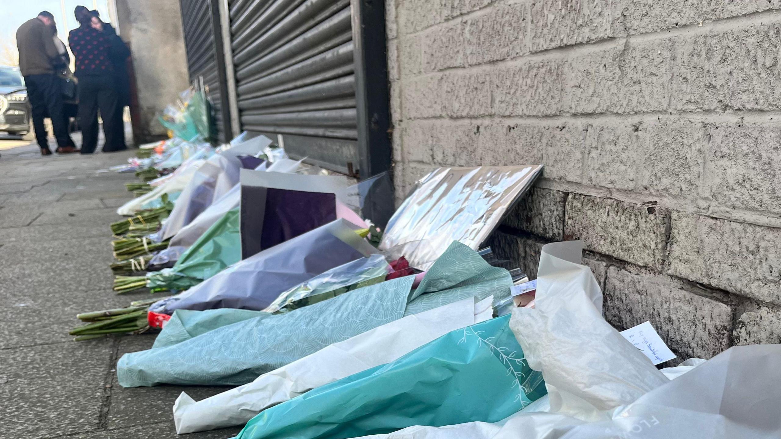 About 20 bouquets of flowers, some with messages attached, in a row on the ground outside a grey, brick-built building with black shutters. In the background, people can be seen standing on the pavement.