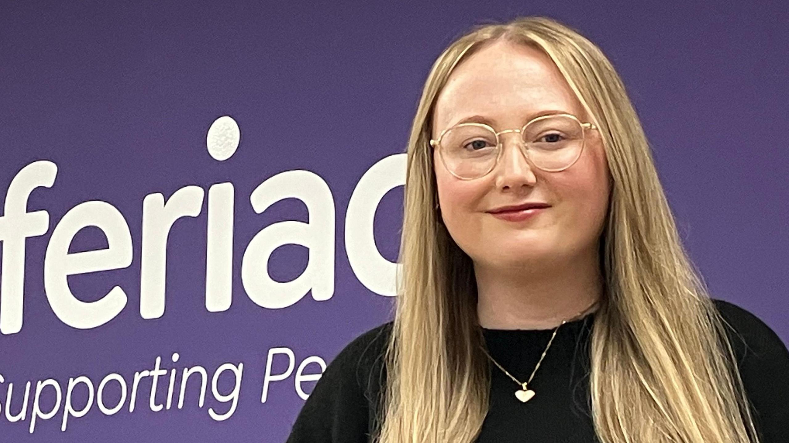 Mae gan Carys Williams wallt hir golau ac mae'n gwisgo sbectol gron a siwmper ddu. Mae Carys yn sefyll o flaen wal borffor gyda logo elusen Adferiad y tu nol iddi.