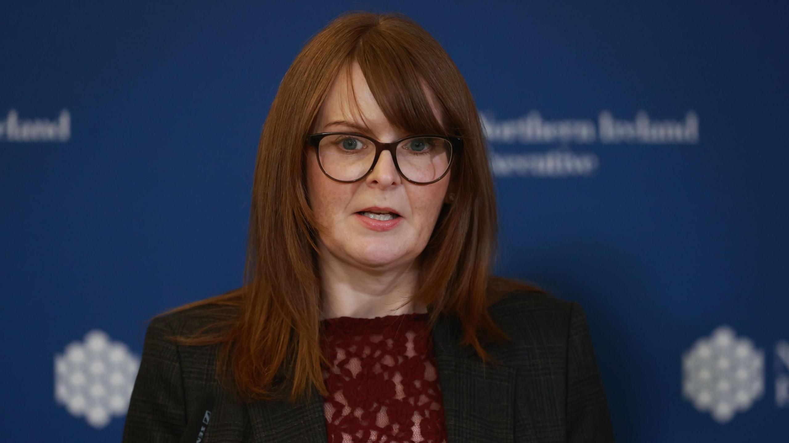 Dr Caoimhe Archibald wearing a dark coloured suit with a blue background behind her. She has long reddish hair with a fringe and has glasses on.