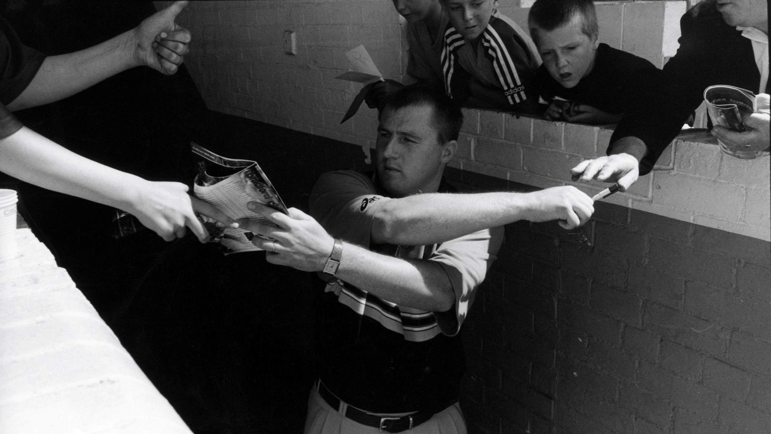 A black-and-white photo of a man in a polo shirt crossing his arms as he tries to sign a football programme while giving back a pen to a young football fan.