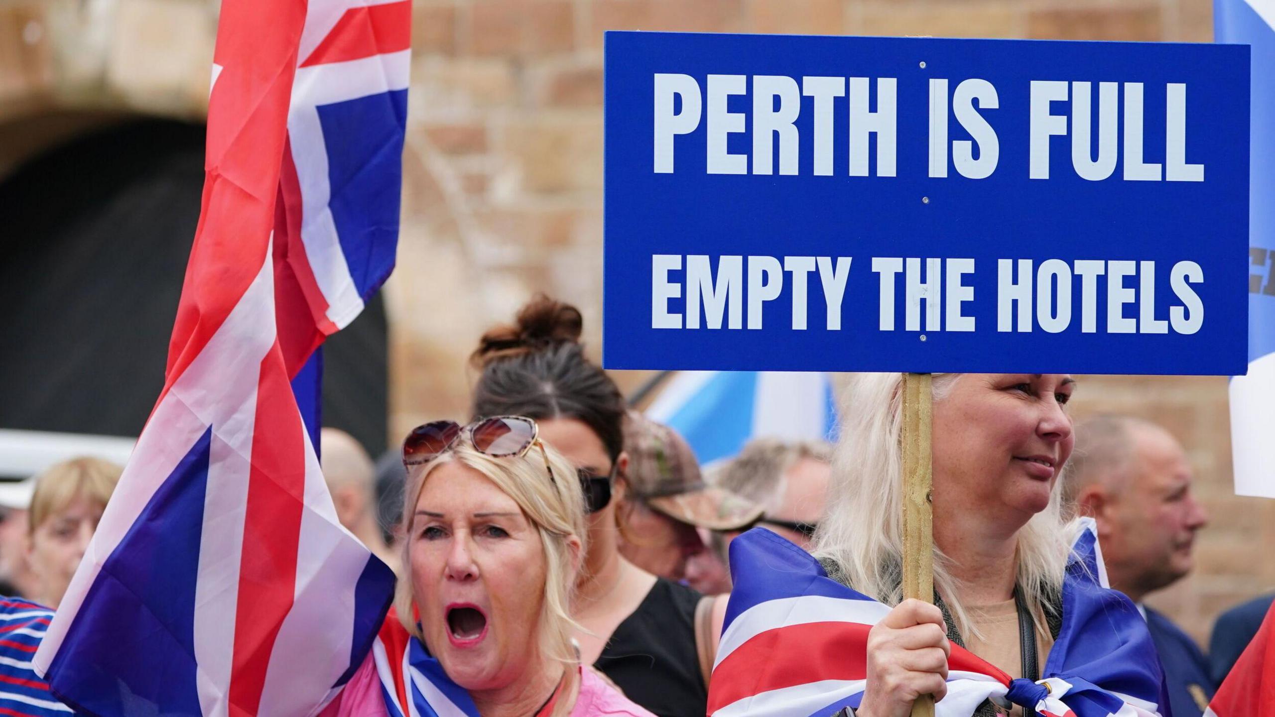 People demonstrating at an Abolish Asylum System protest outside the Radisson Hotel in Perth. Two blonde women are wrapped in Union Jack flags and one holds a sign which reads "Perth is full empty the hotels"