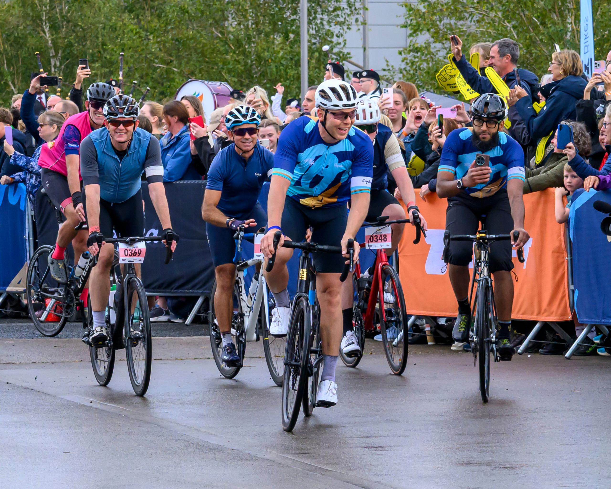 Four cyclists in lycra, helmets and sunglasses, cycling on a smooth road surface, led by Sir Chris Hoy. There are large crowds behind barriers. 