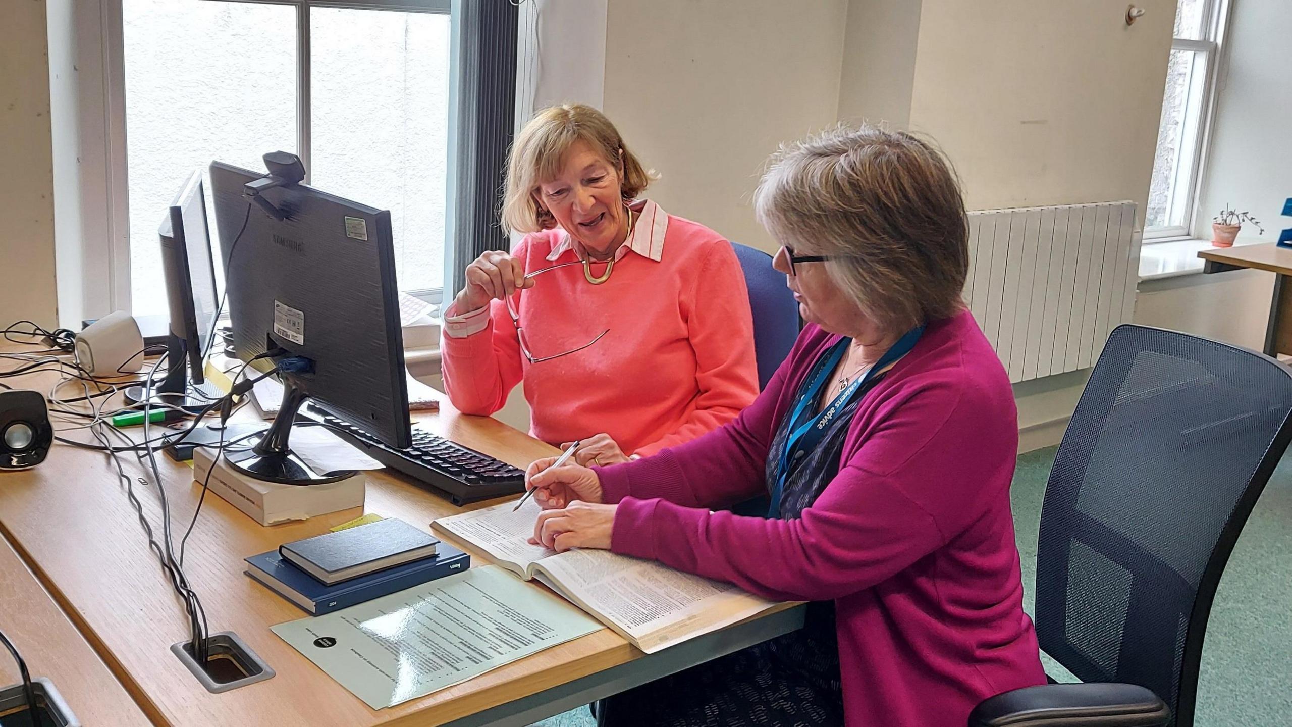 Two women in front of a computer screen looking at book on a desk. One is wearing a coral jumper and holding her glasses. The other has a blue dress and pink cardigan and is holding a pen. 