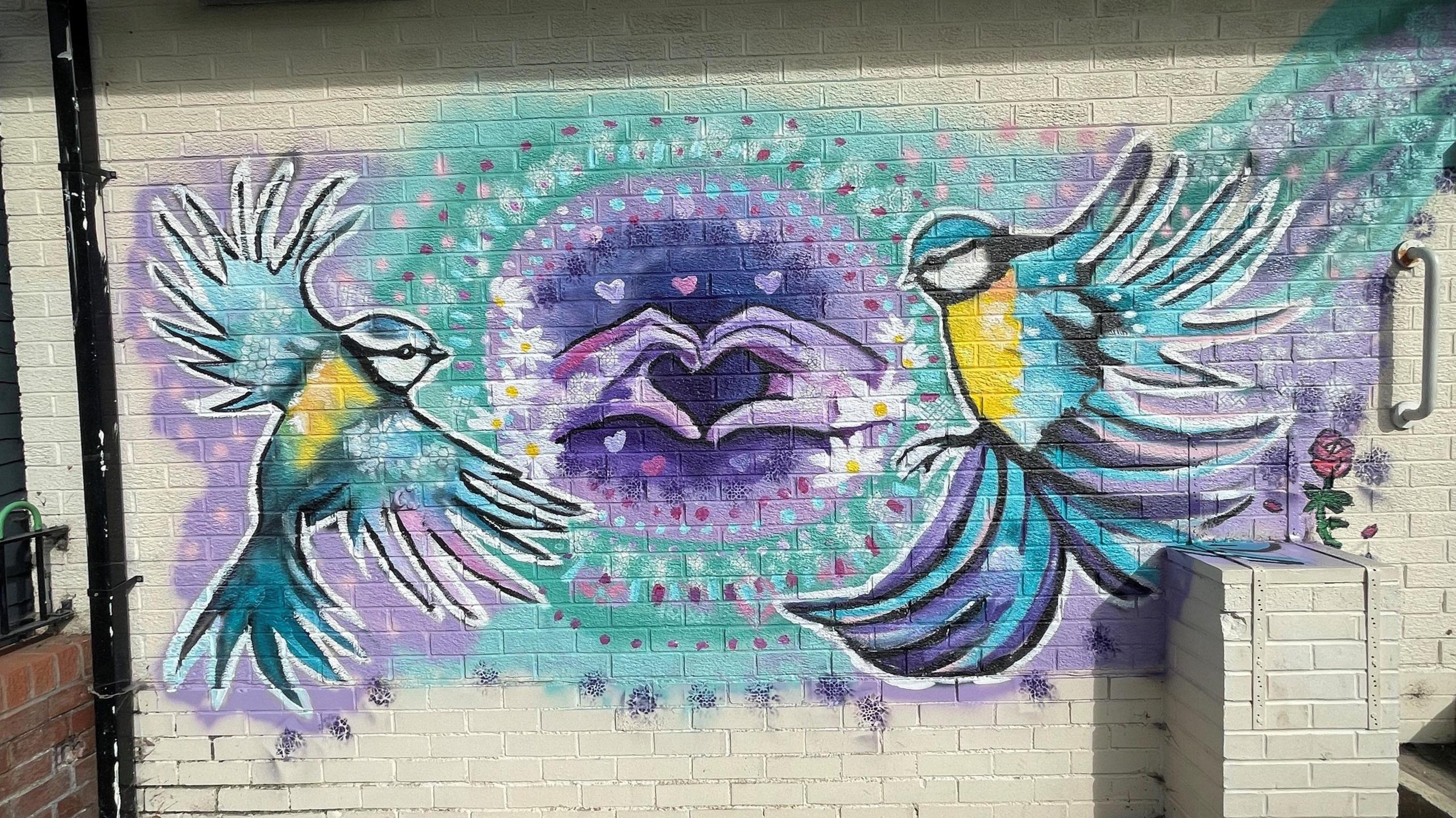 A mural showing two birds with yellow chests, blue wings and white faces against a blue and purple background. The image also features two hands making a heart shape with their fingers in the centre of the artwork. 