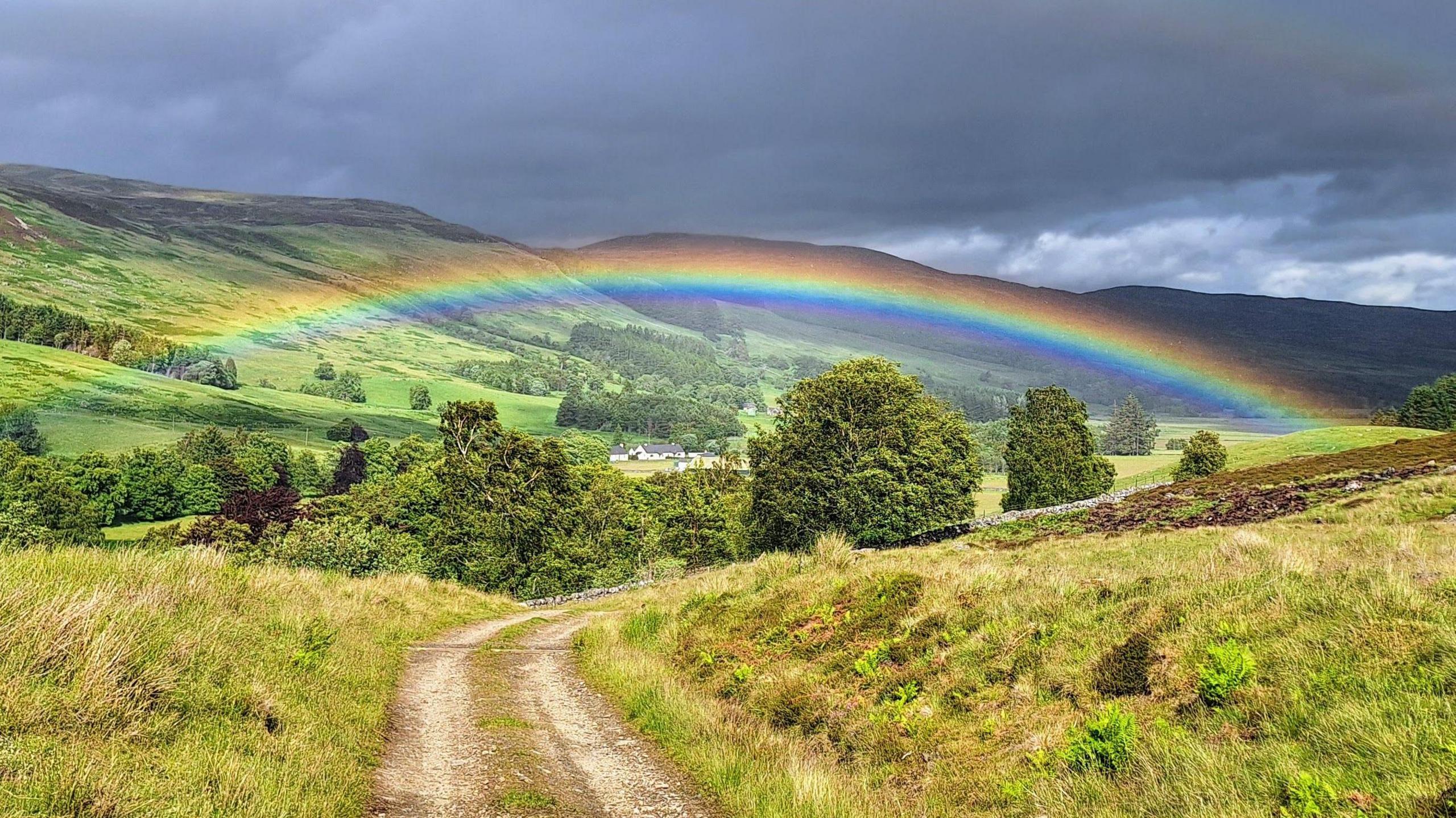 A rainbow with a number of colours over a dirt track surrounded by grass verges leading towards a cluster of trees. Beyond the trees is at least one building, which is brightly-coloured. The sky is a mixture of dark grey colours.