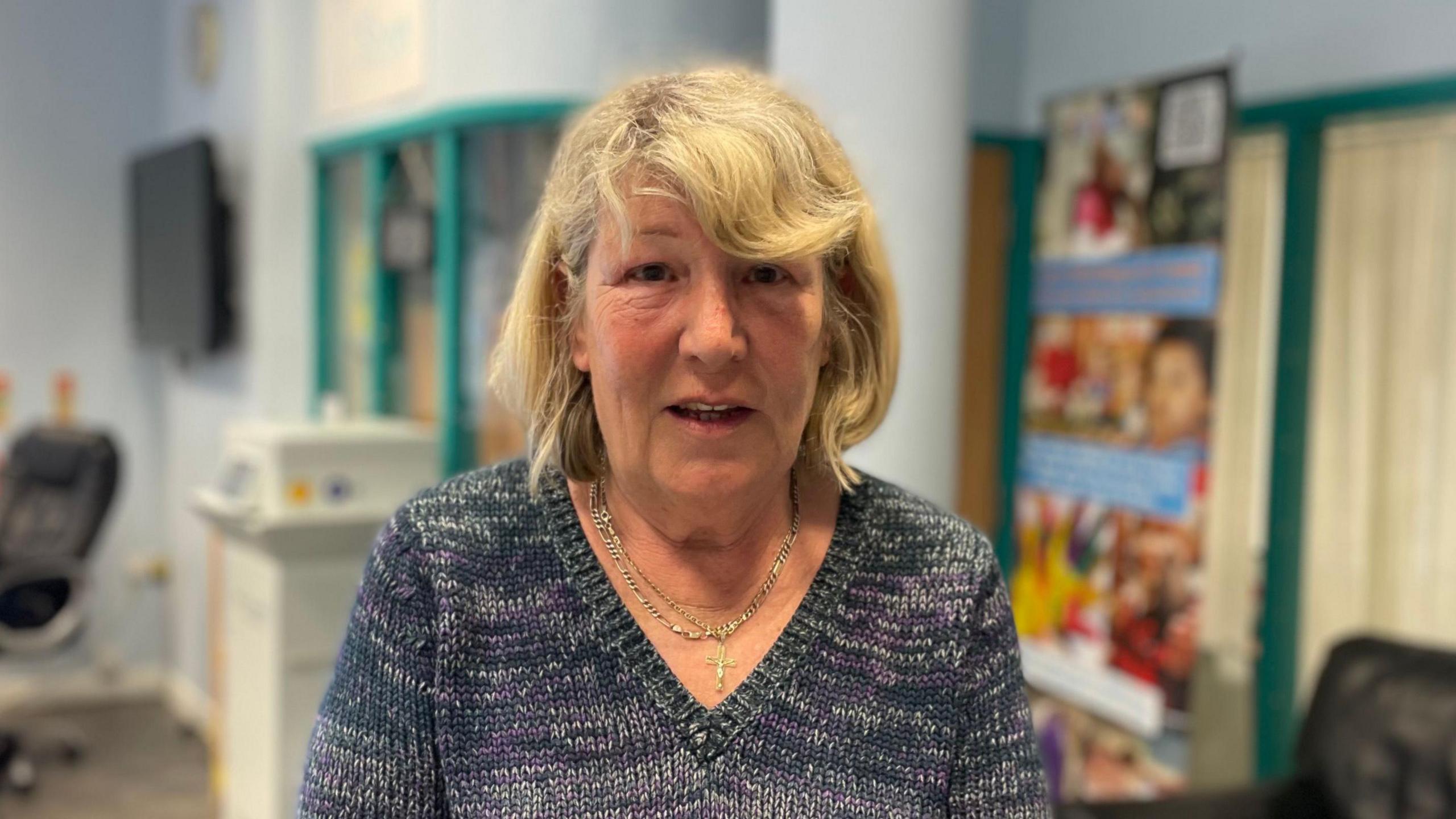Anita Grodkiewicz looking at the camera. In the background is the community centre lobby, with chairs and an information board. She has shoulder-length blonde hair and is wearing a blue and grey V-neck jumper.