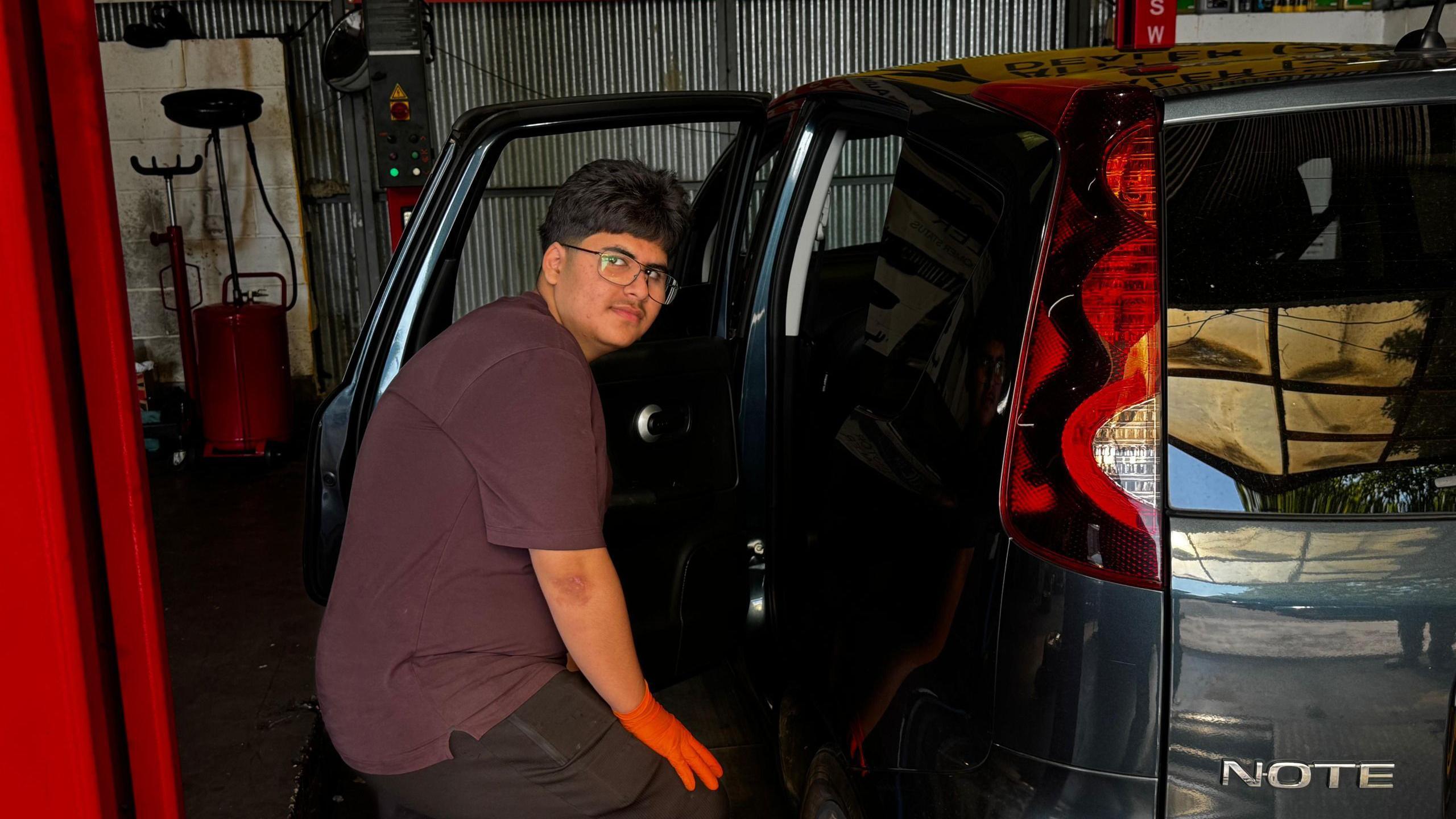 Azhe is seen wearing orange gloves as works on a dark-coloured car inside a vehicle workshop. The rear door is open, with garage equipment and automotive service banners visible in the background.
