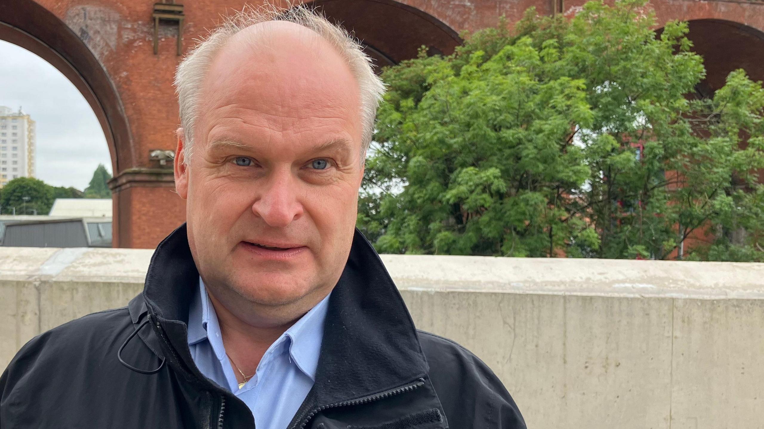 Michael Handley, who is wearing a blue shirt and black coat, stands by a wall with Stockport viaduct's arches seen behind him.