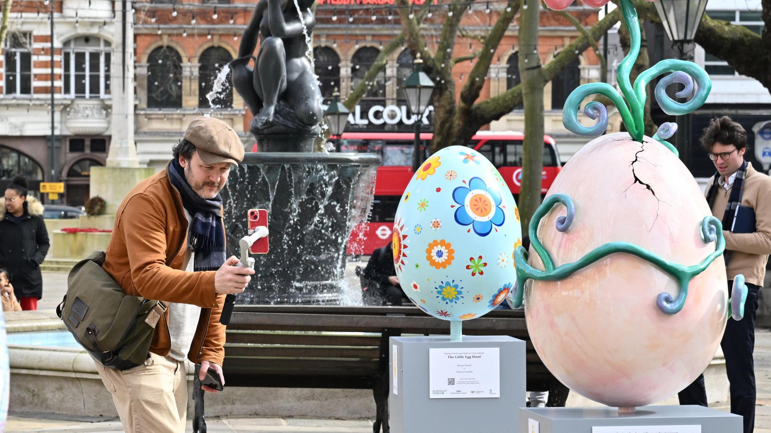 A man taking a photo of decorated eggs, one is blue with funky flowers and the other looks cracked with a flower with an eye in the middle coming out of the top. 