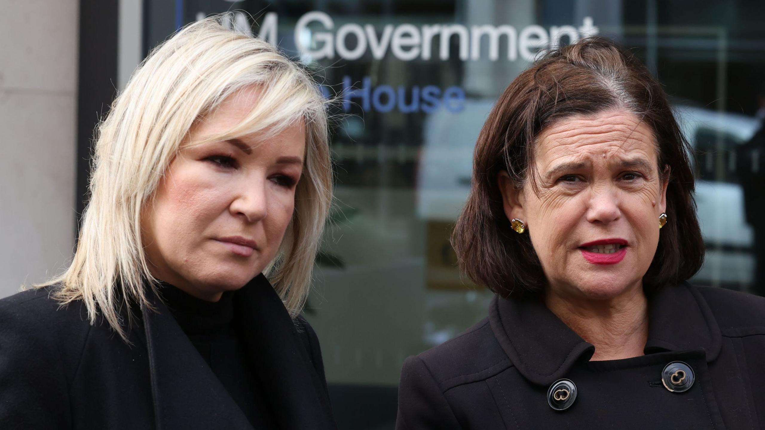 Sinn Féin deputy leader Michelle O'Neill (left) and leader Mary Lou McDonald (right). O'Neill has blonde shoulder-length hair and McDonald has a dark brown bob. Both are wearing black coats.