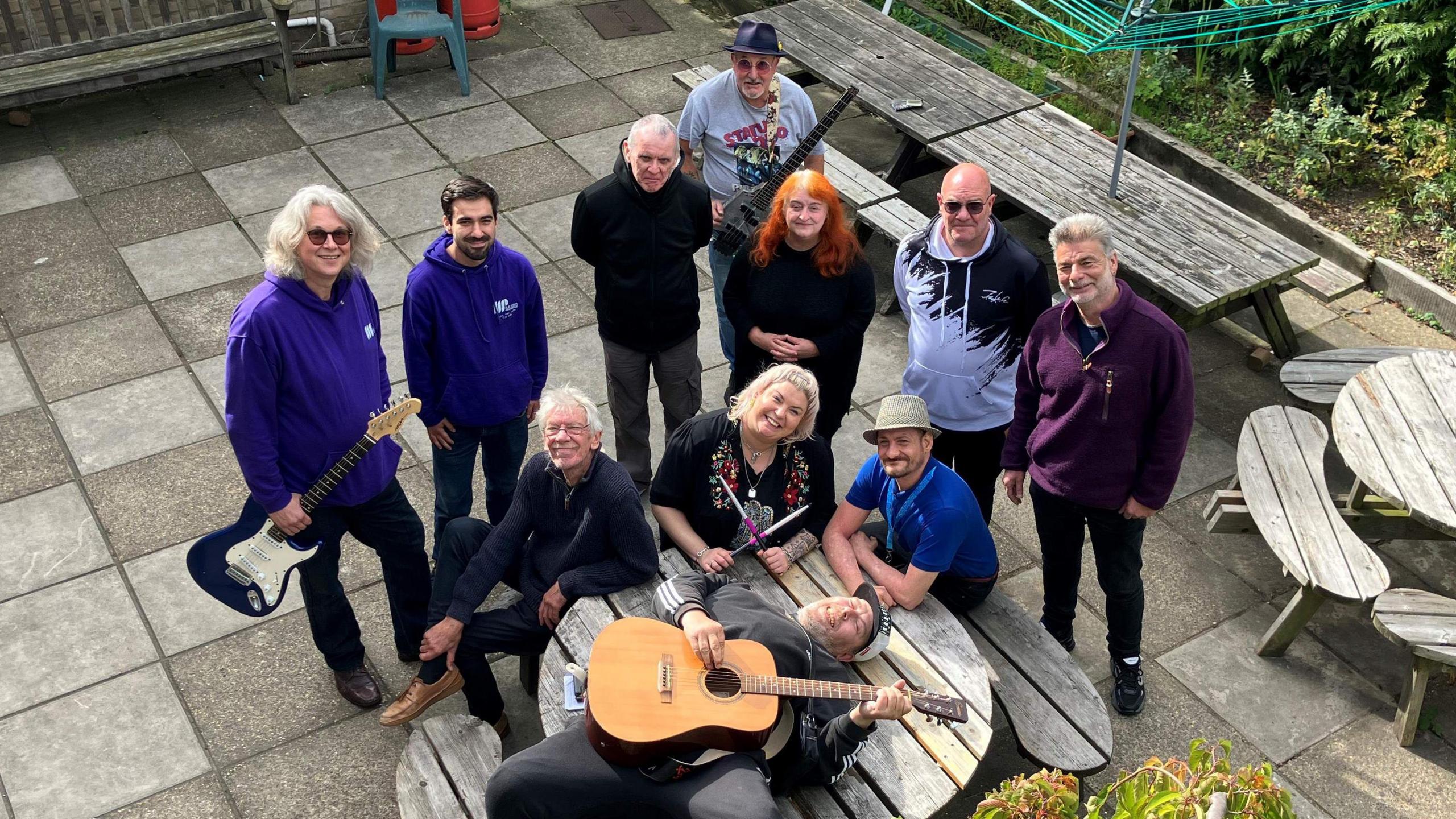 11 people gathered in a courtyard around a picnic bench, looking up towards the camera. One man is lying on the table holding a guitar