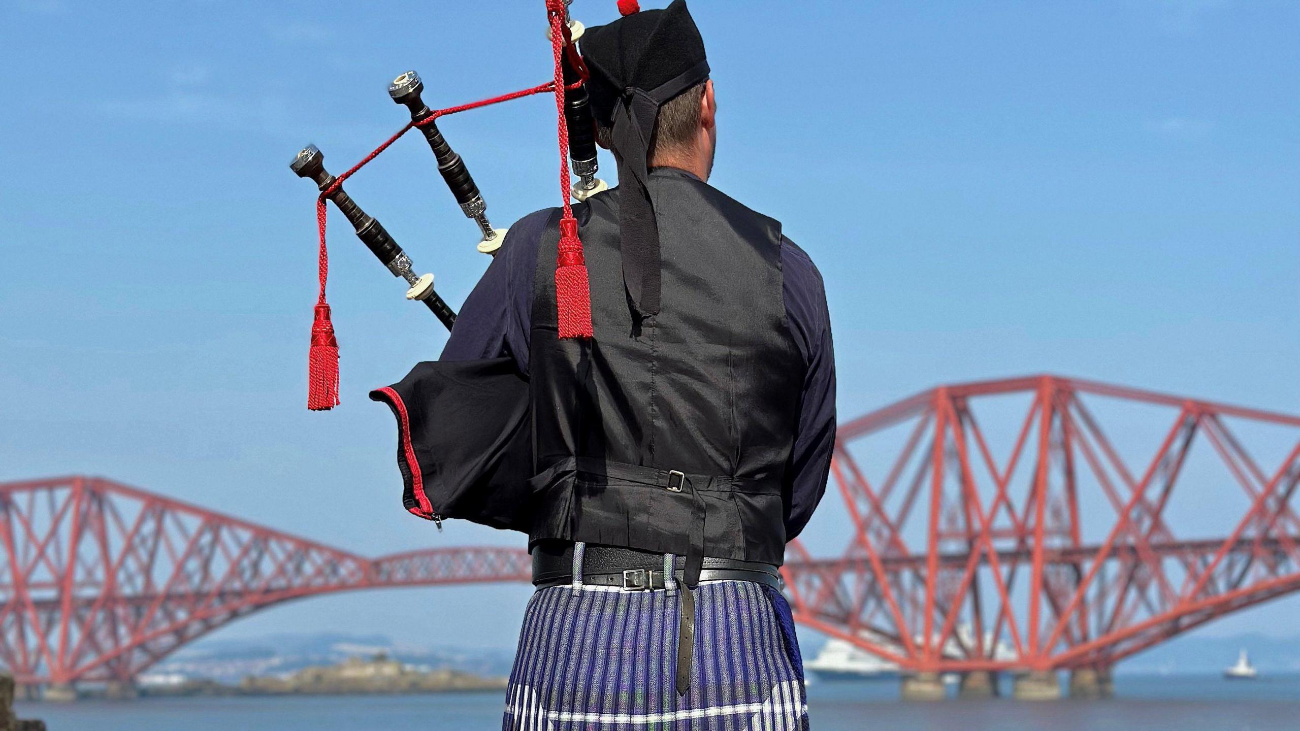 A bagpiper, wearing a full kilt playing his instrument in front of the Forth Bridge.