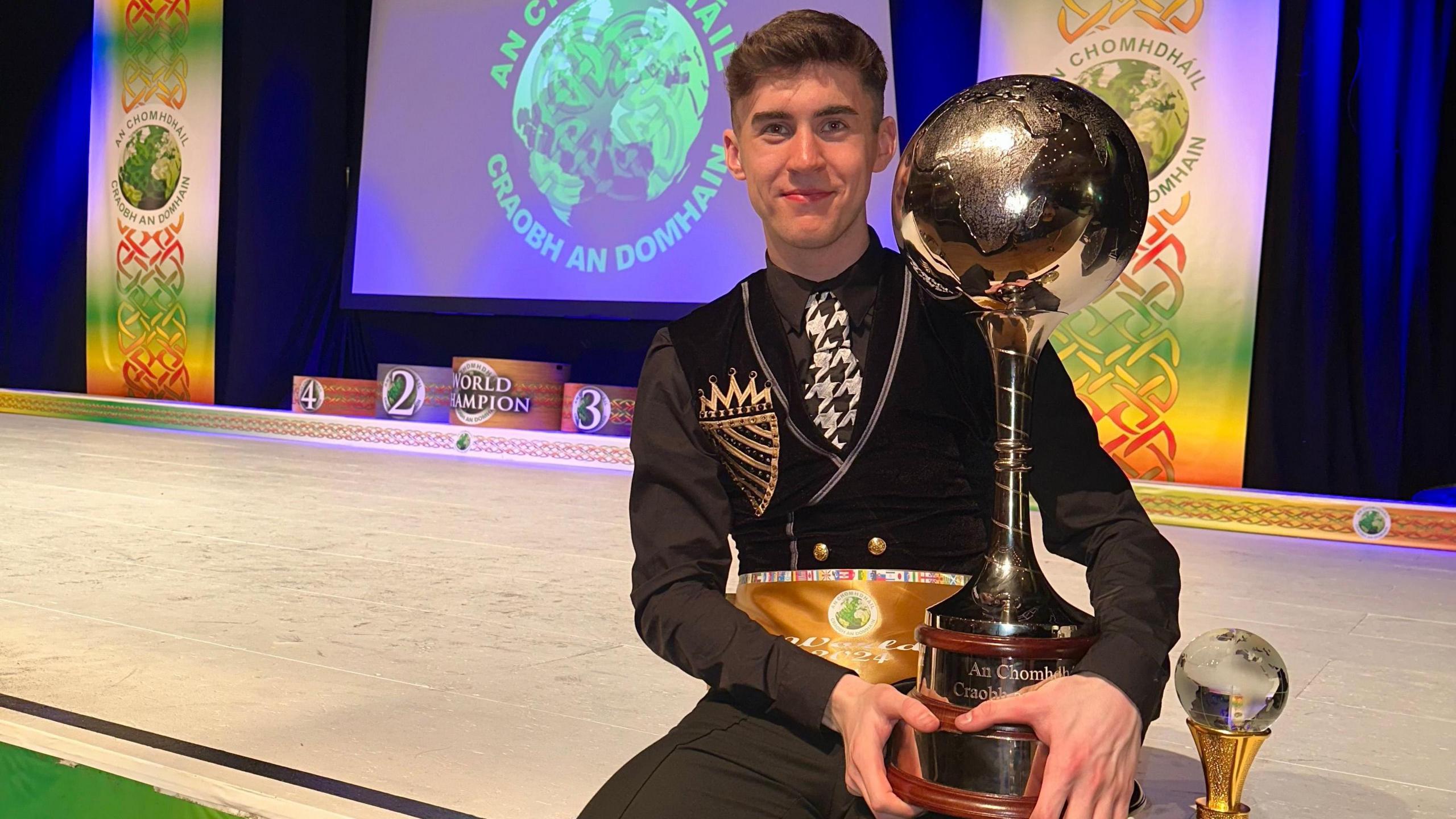 Barry, with dark short hair, smiles at the camera as he holds a large silver trophy. He is sitting on a stage with a projector screen behind him. He is wearing a black shirt and trousers, a black waistcoat with gold button designs and a patch and a white, grey and black tie.