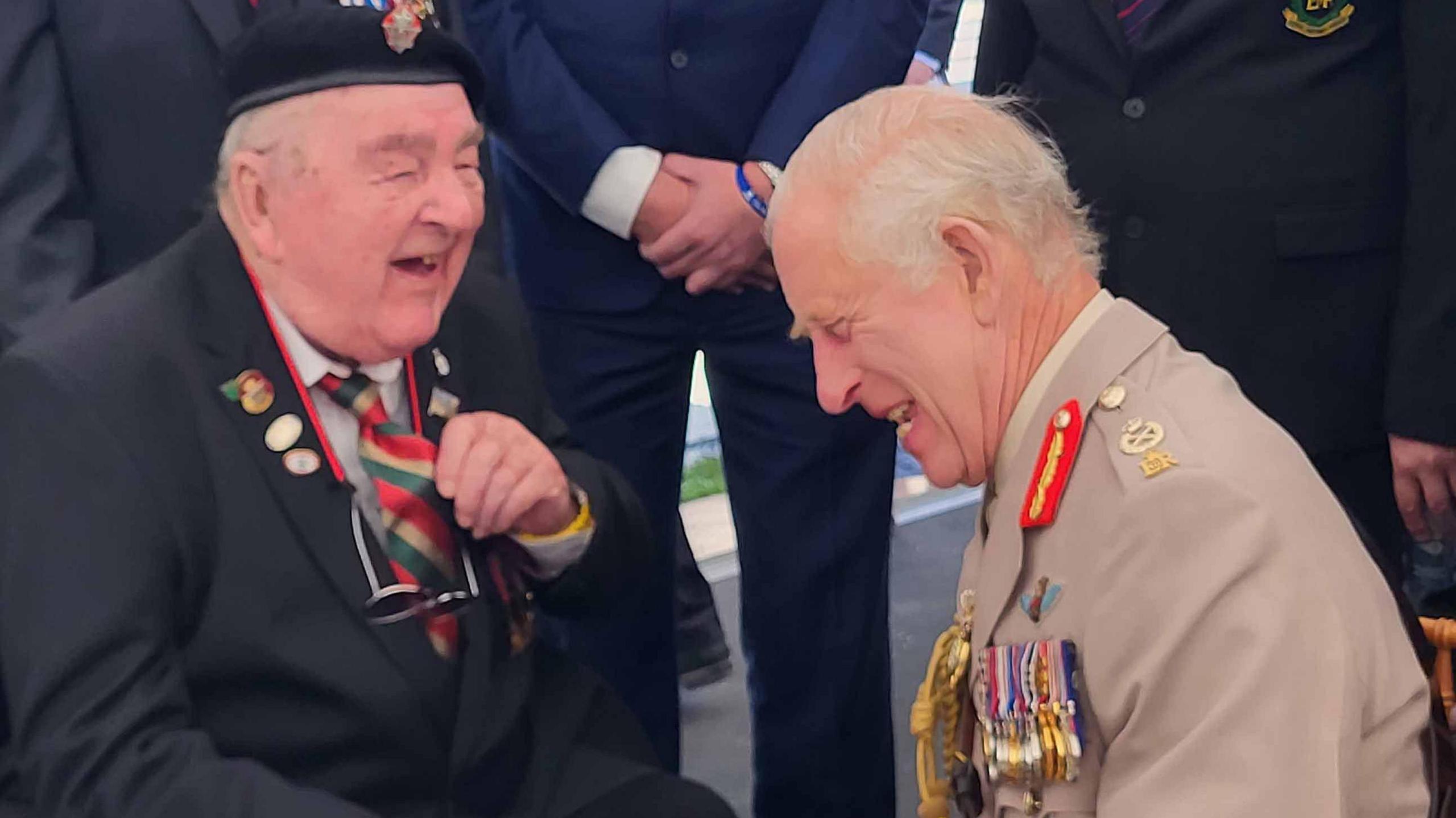 Richard Aldred is sitting down facing King Charles III. The King has his head down and eyes shut as he laughts. Mr Aldred is looking at him and laughing as well. Both are wearing their military uniforms which are adorned with medals, badges and epaulets.