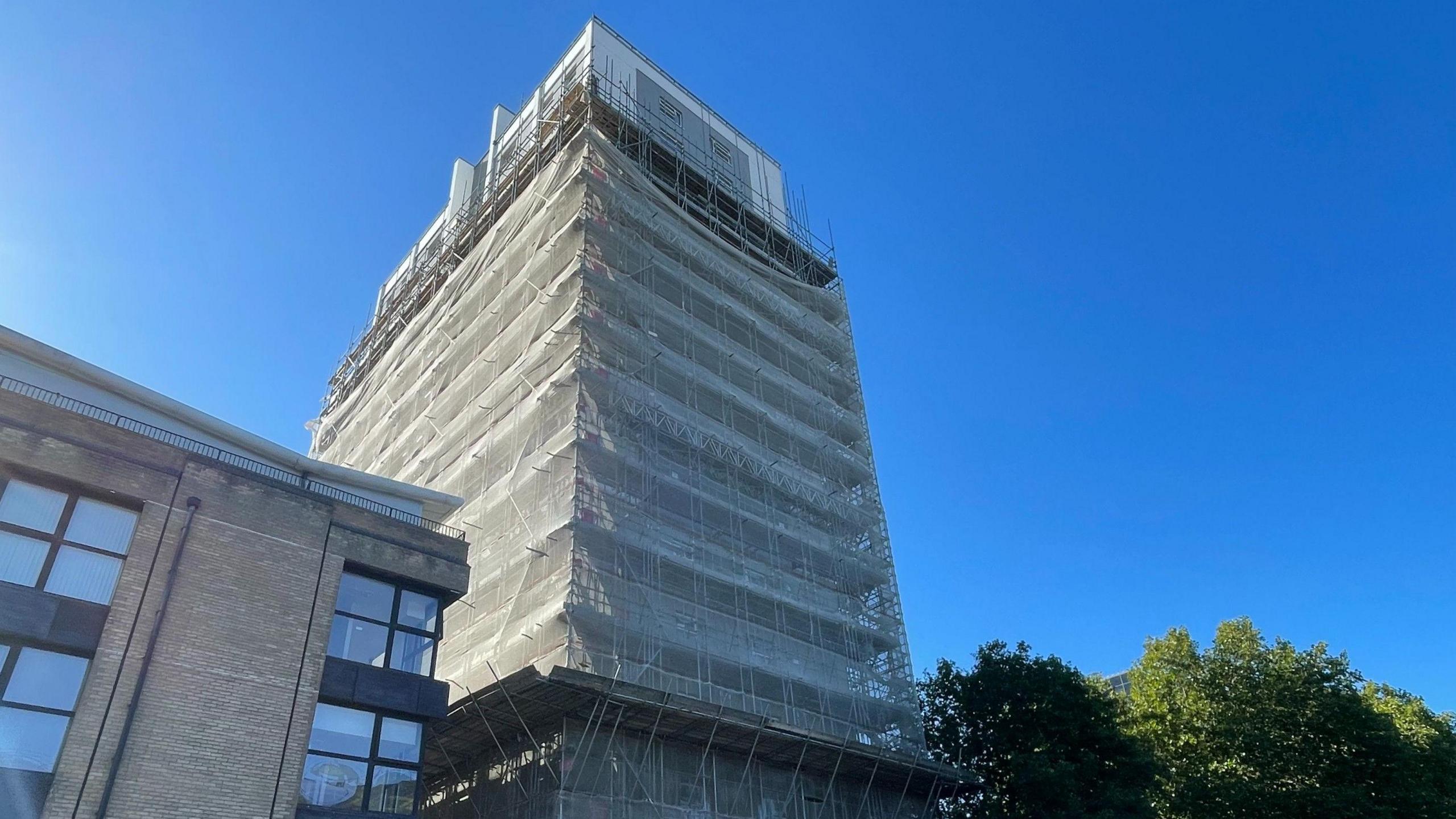 St Francis Tower, most of the tower is covered in scaffolding and netting, the top portion of the block is no covered in scaffolding and has a cladded finish. There is a bright blue sky behind with green trees to the right. On the left is a brick and glass building.