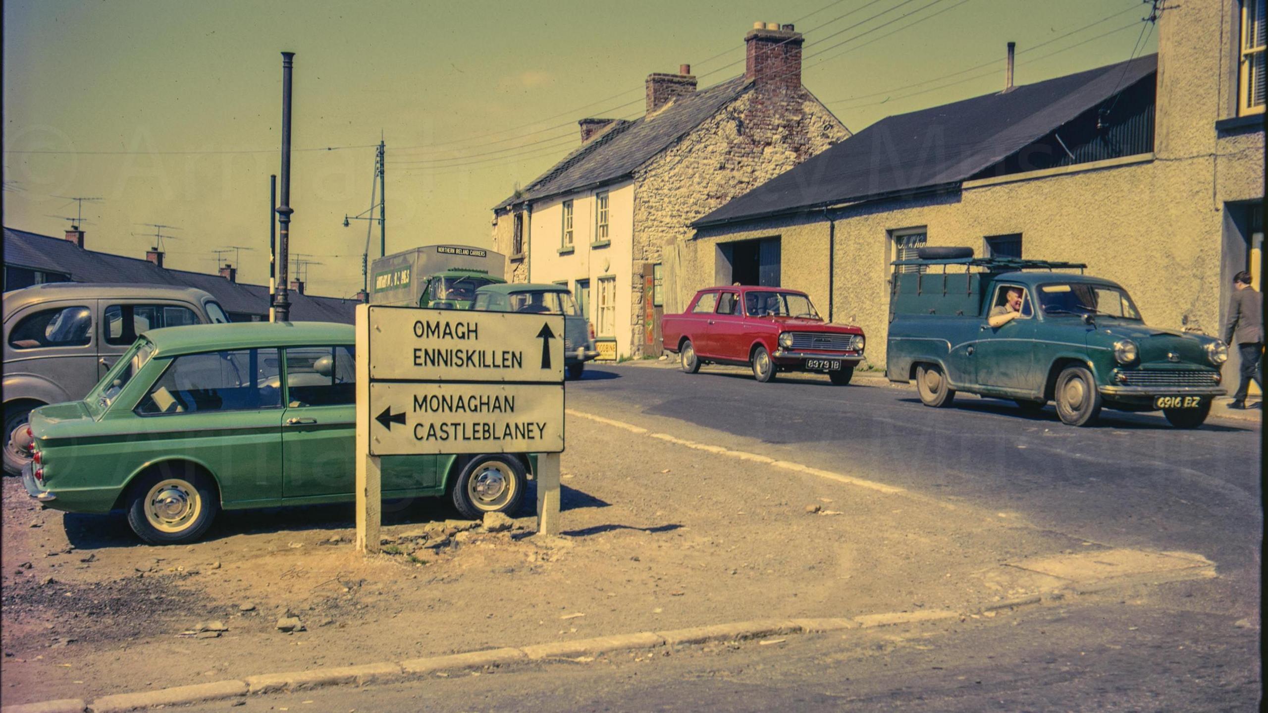 A digitalised photo from a film of street signs. Green, red and blue cars can be seen parked along the side of a small street. Looking west from Irish Street corner several cars are at the junction near the camera and the roofs of the terrace of new houses a partially visible in the background. An old and partially derelict house is on the right of the frame and a road sign reads "Omagh Enniskillen, Monaghan Castleblaney". 