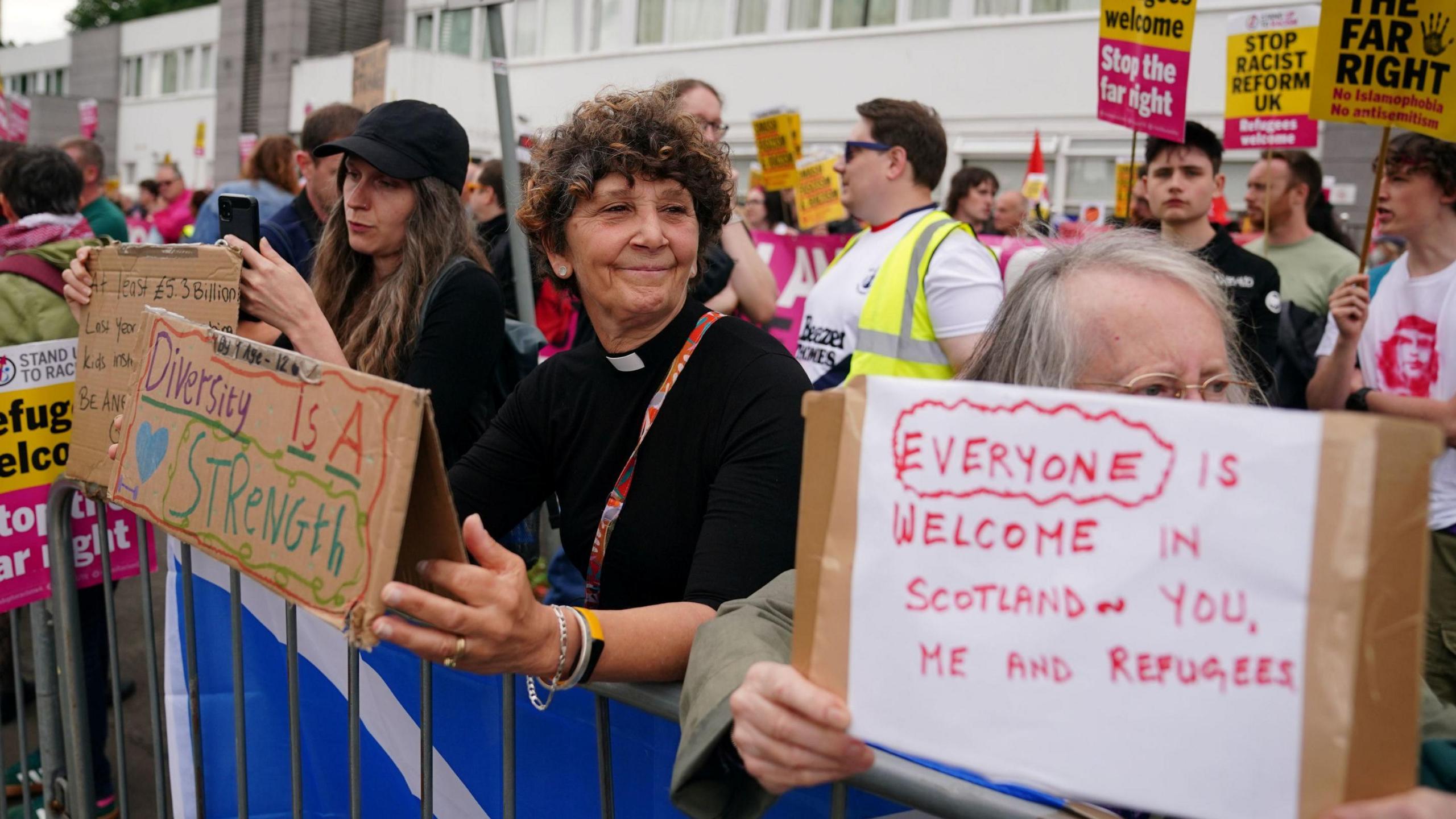 A group of people stand behind a metal barrier with messages supporting diversity and refugees.