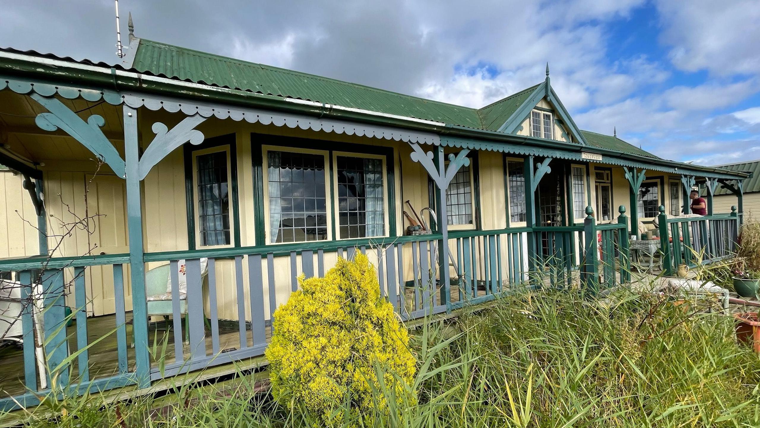 The outside of Towerview, an early 20th Century prefabricated bunglow. It has a green tin roof and wooden walls painted cream. The roof just out across a veranda, which has a wooden balustrade. In front of the house are reeds