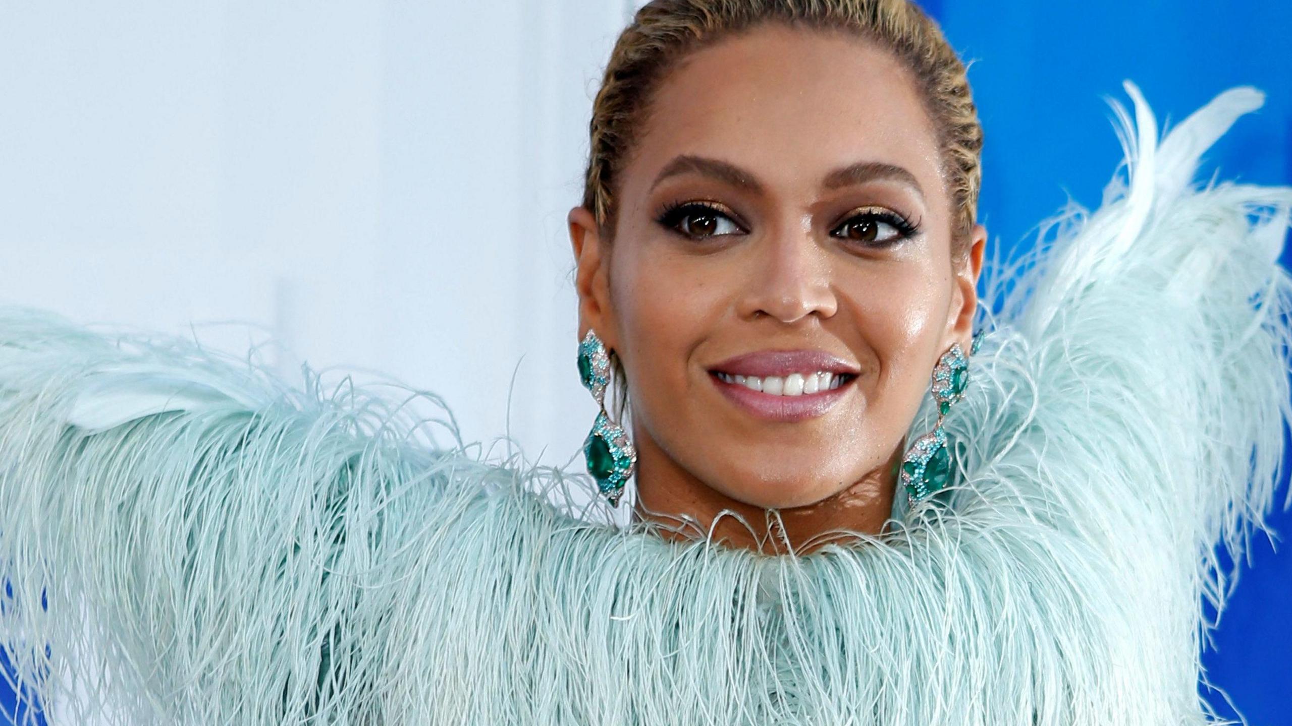 Beyoncé at a reception, wearing pale blue ostrich feathers and giant turquoise gemstone earrings. She is smiling against a mid-blue background. 