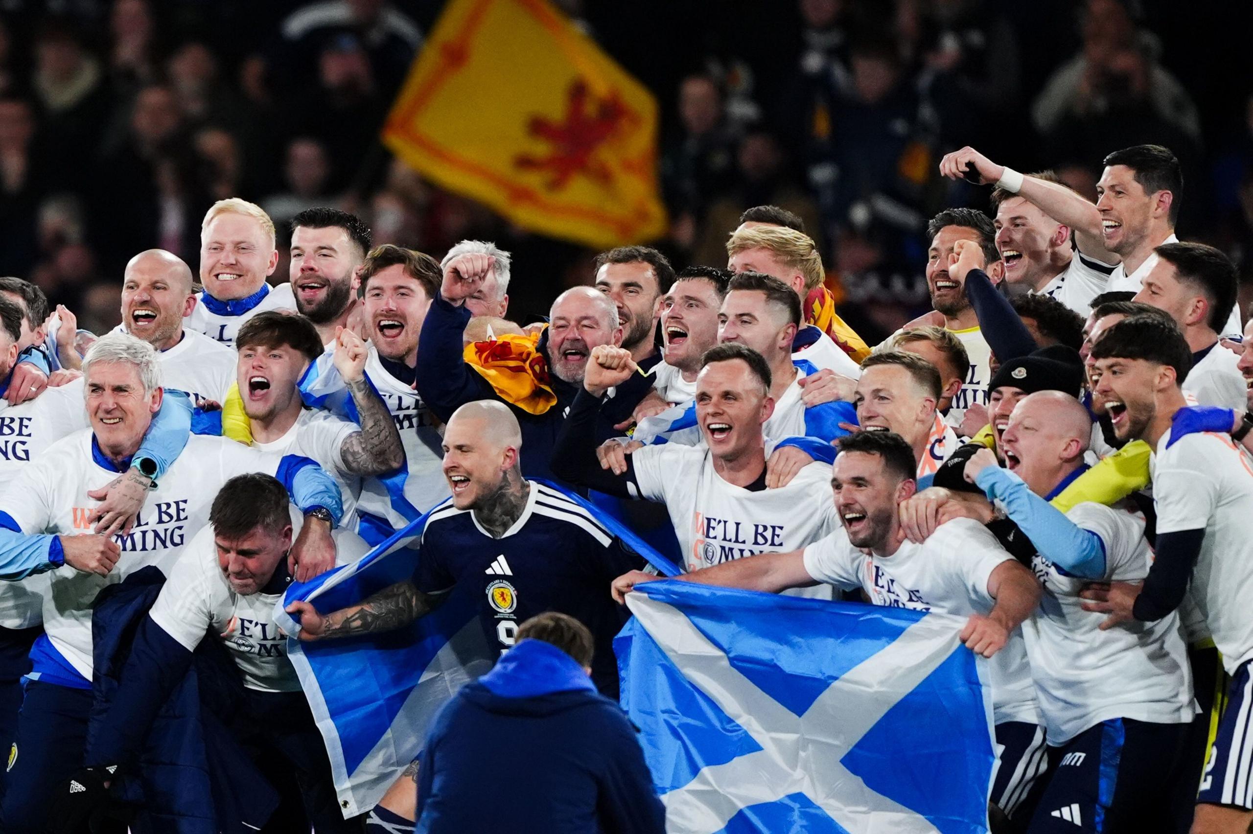 The Scotland's men's football team gather together for a group photo after winning their match against Denmark. They are wearing tops which say "We'll be coming" and holding blue and white Scotland flags. Behind them, fans can be seen in the stands waving a yellow and red Lion Rampant flag.