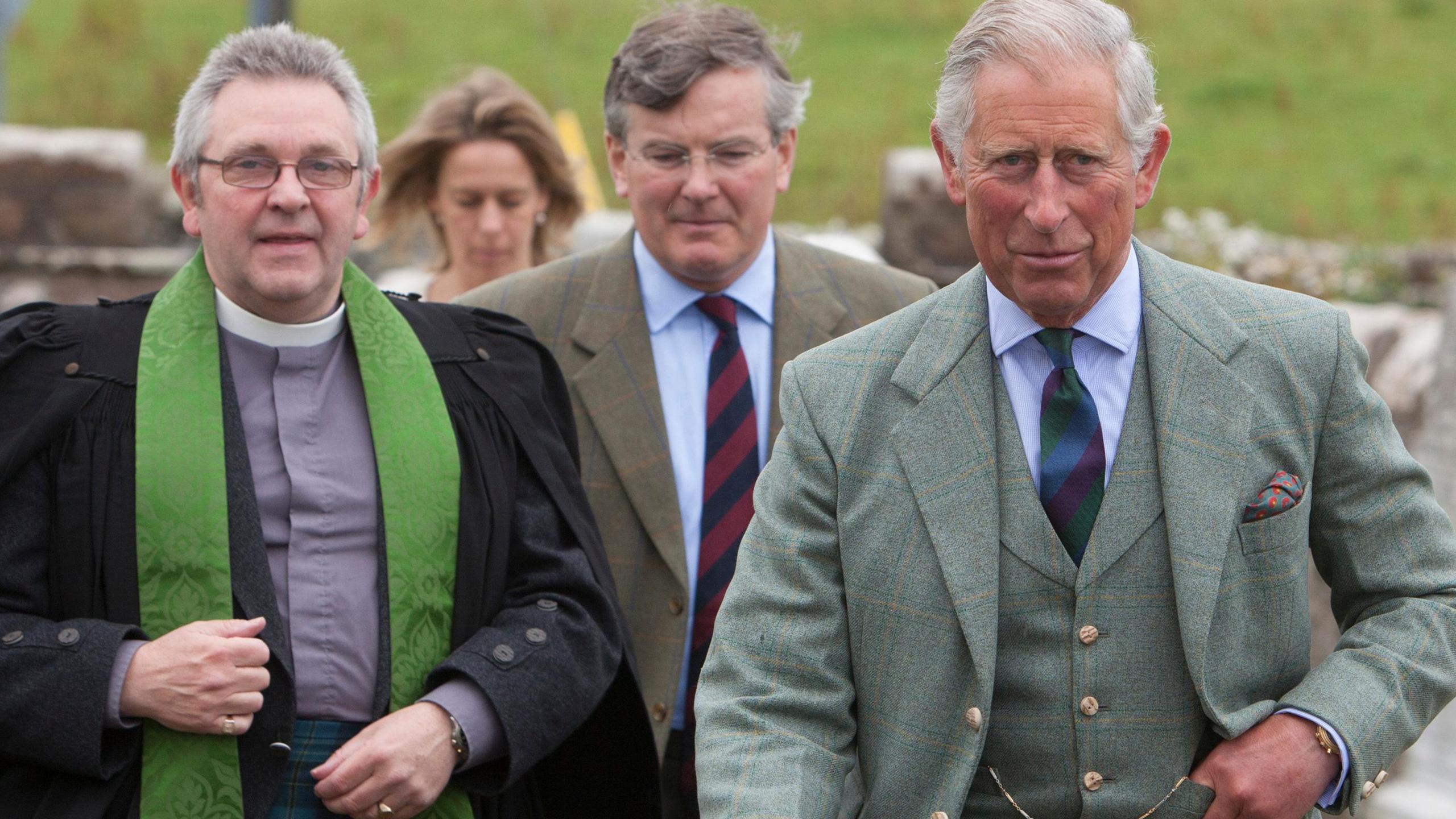 The Reverend Ivan Warwick walking in a churchyard with King Charles. The minister is wearing a white clerical collar and a grey shirt. He has a long green clerical shawl and a black cloak. The king is wearing a kilt and brown leather sporran. He as a waistcoast and jacket in matching patterns.