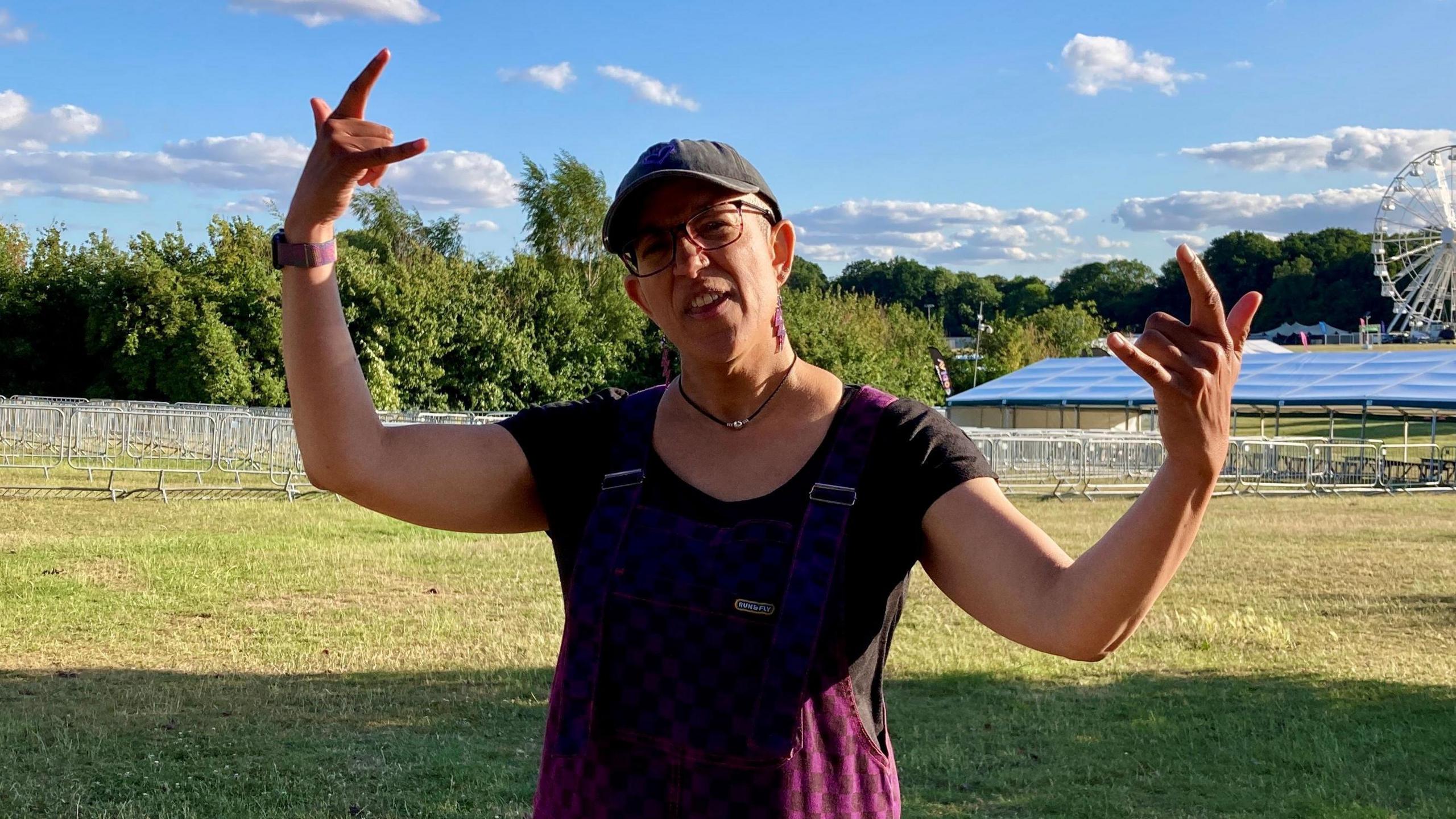 A woman standing in an empty field at a festival. She is wearing a dark purple top and a cap. She is holding both of her arms up and using her fingers sign the "rock on" symbol, with her thumb, index finger and little finger up. There is a white big wheel in the background and the sky is blue.