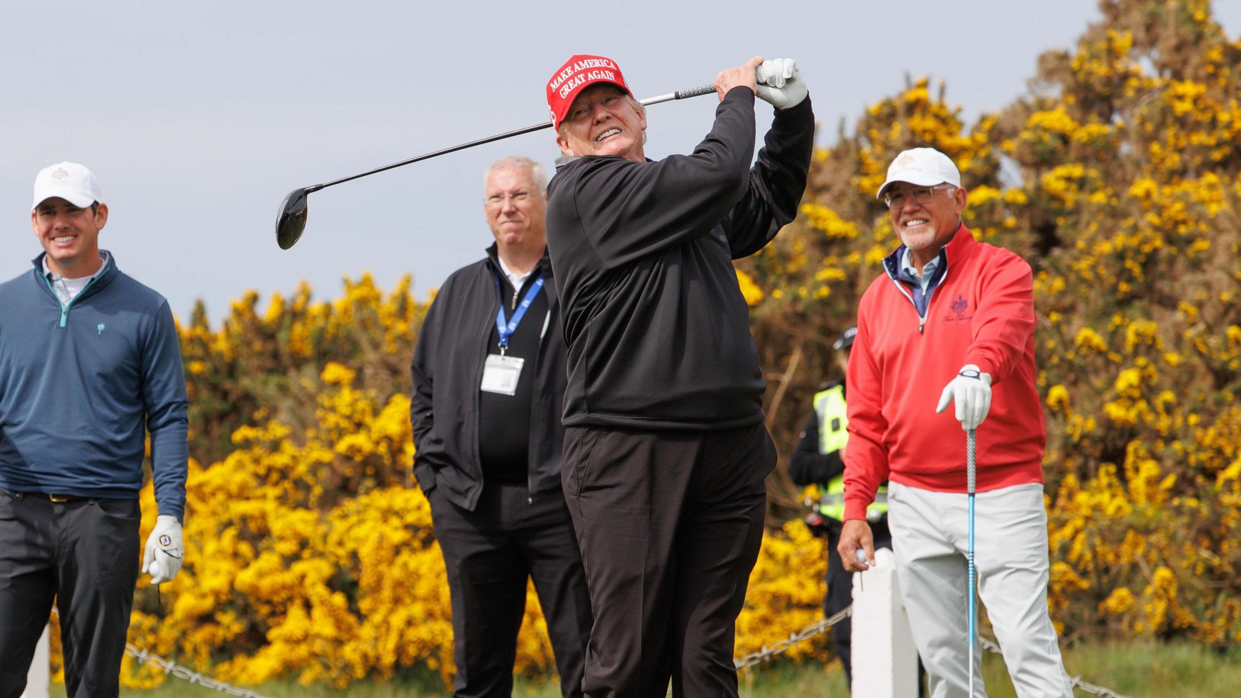 Trump in a maga hat and black golf clothes swings a driver