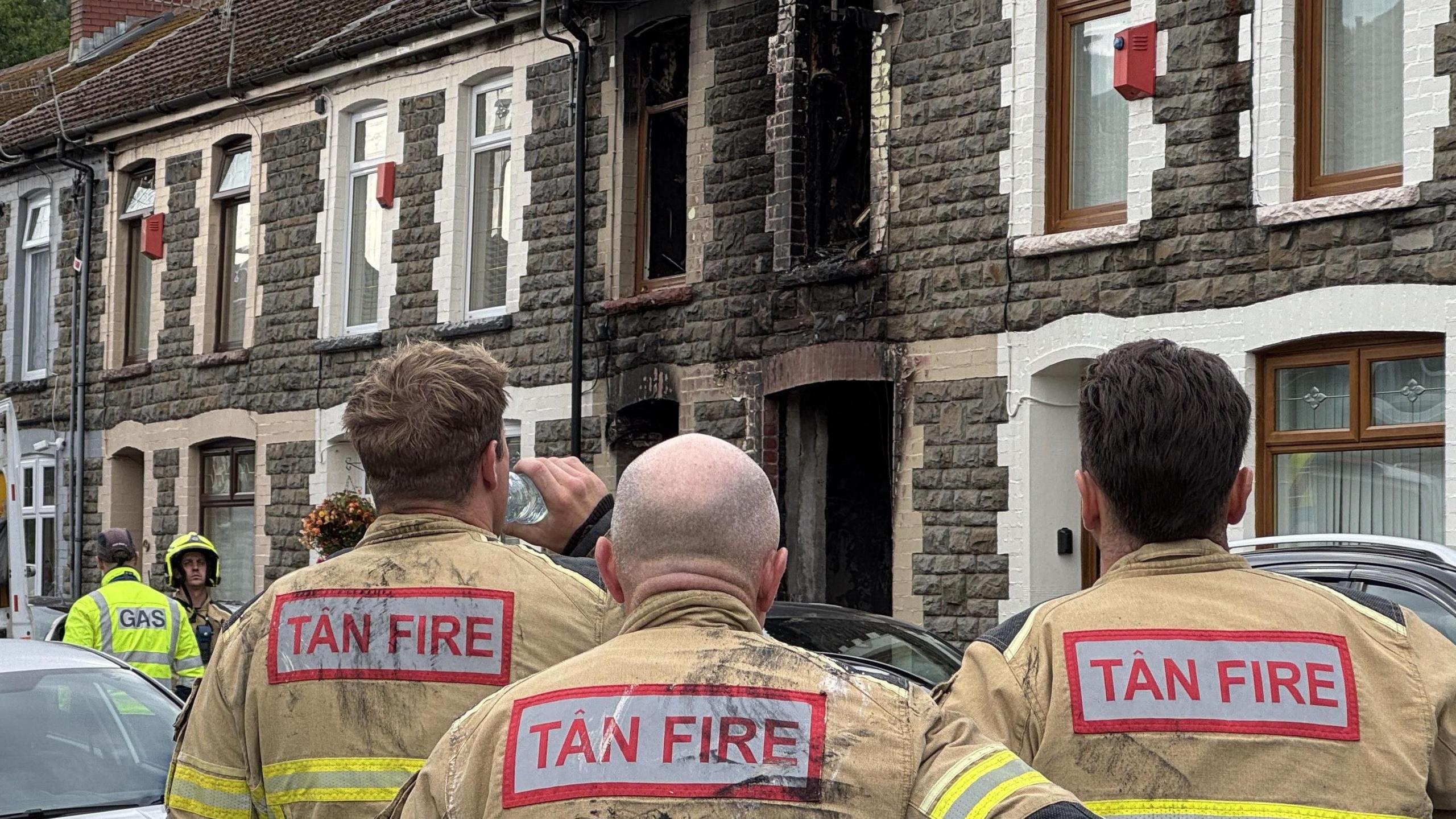 A stone terraced house with fire damage around the windows and doors. Three fire officers look at the house with their backs to the camera.