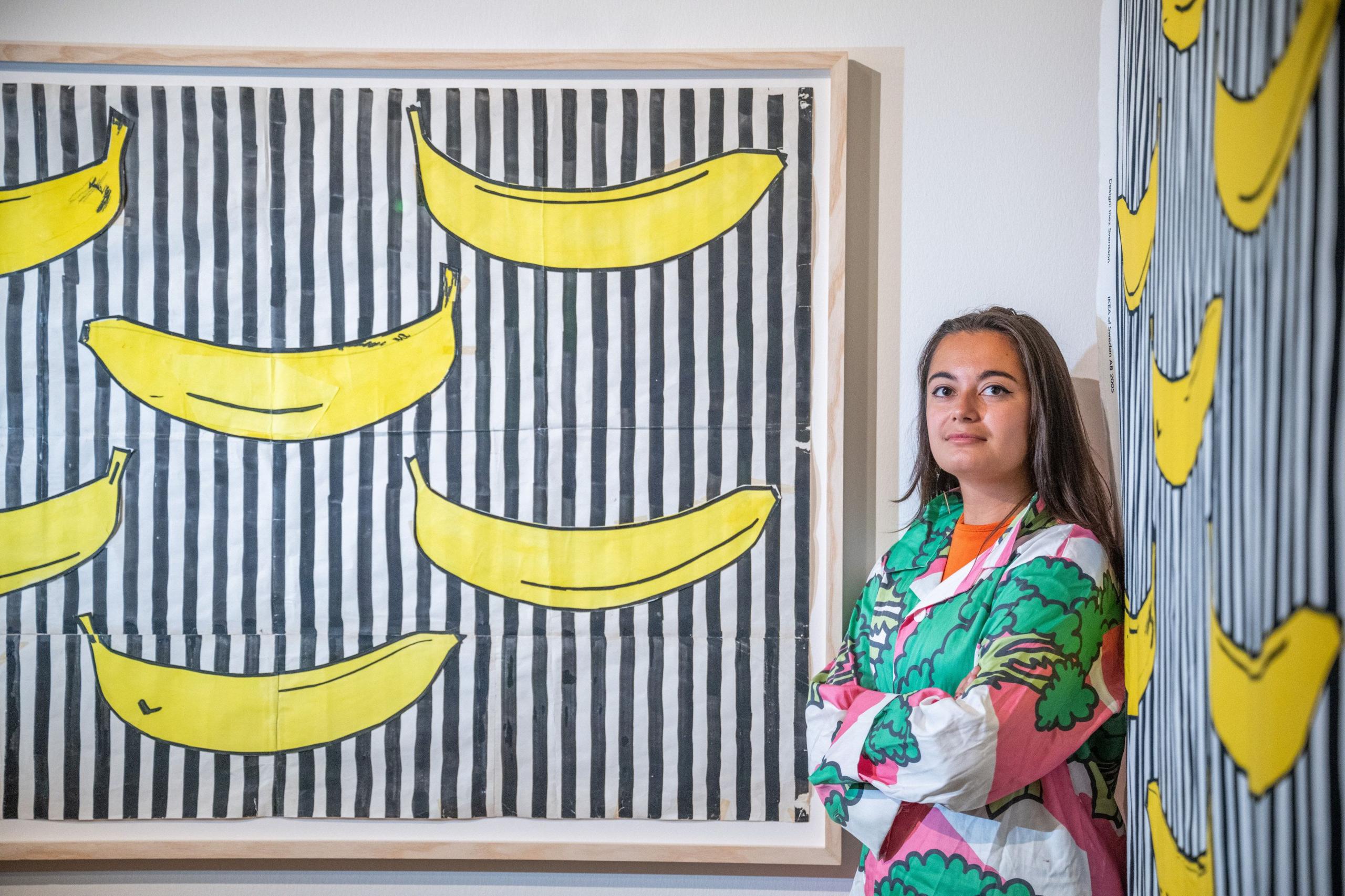 A young woman stands wearing one of the broccoli shirts as she leans against the Randig Banan patterns on two walls - it is a bold black and white striped pattern with large, cartoon bright yellow bananas