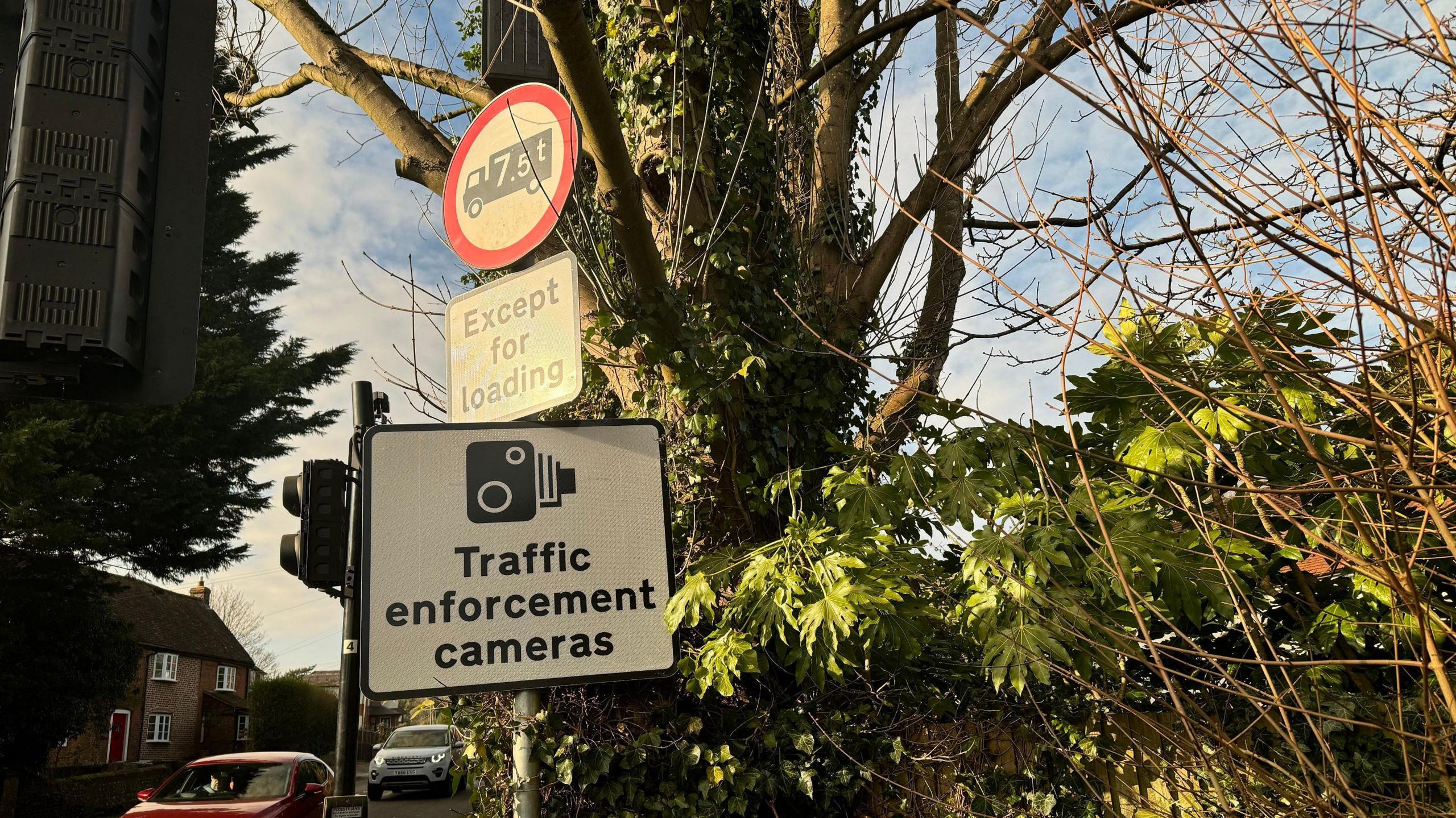 Three signs, one a circle with a red edge which shows vehicles weighing more than 7.5 tonnes are banned, another which says "except for loading", and a sign warning of traffic cameras. In the background, there is a tree and plants. Cars approach a set of traffic lights.