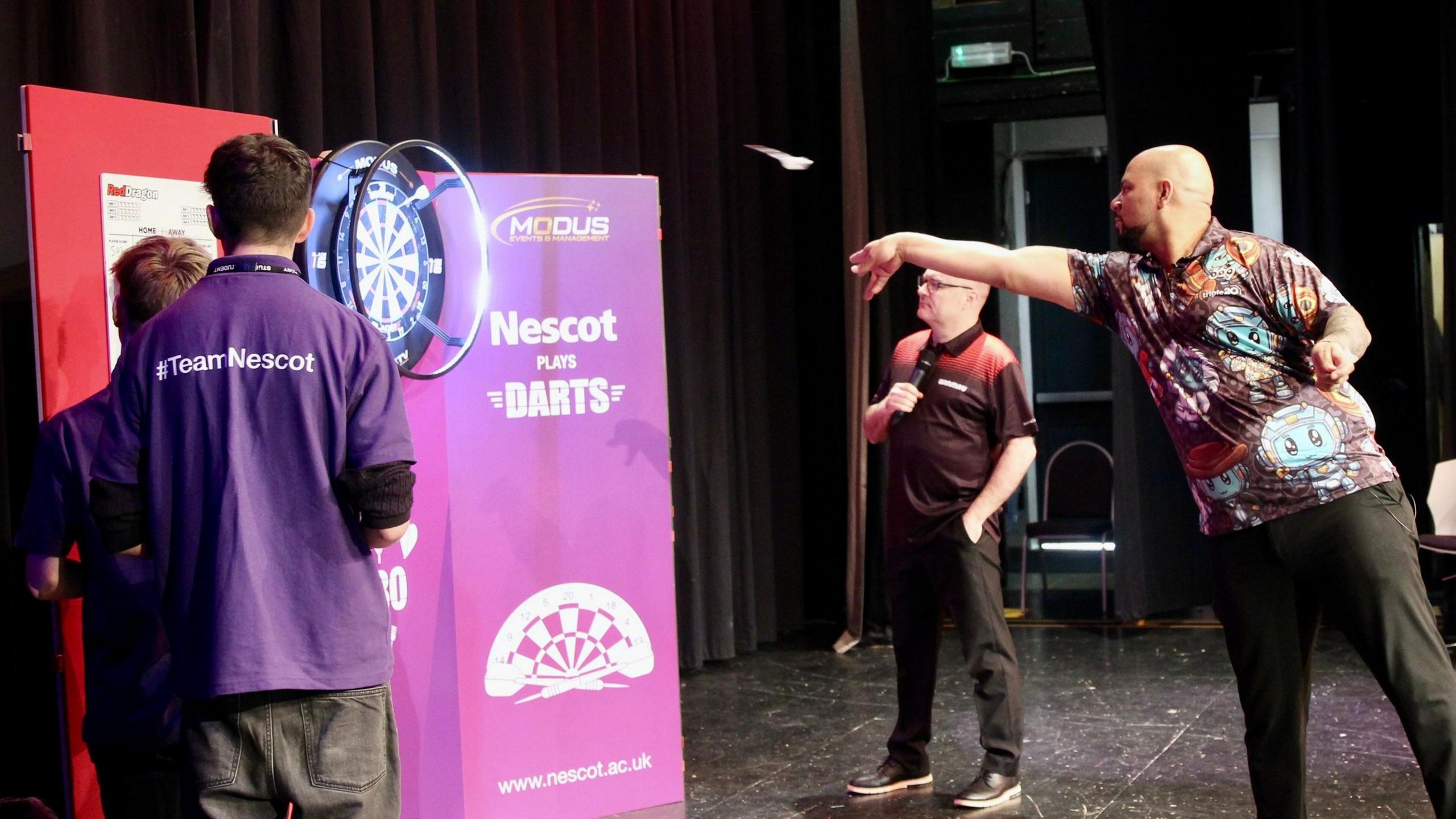 Professional darts player Devon Petersen throws a dart at a board in front of pupils from Nescot who have their backs turned to camera 