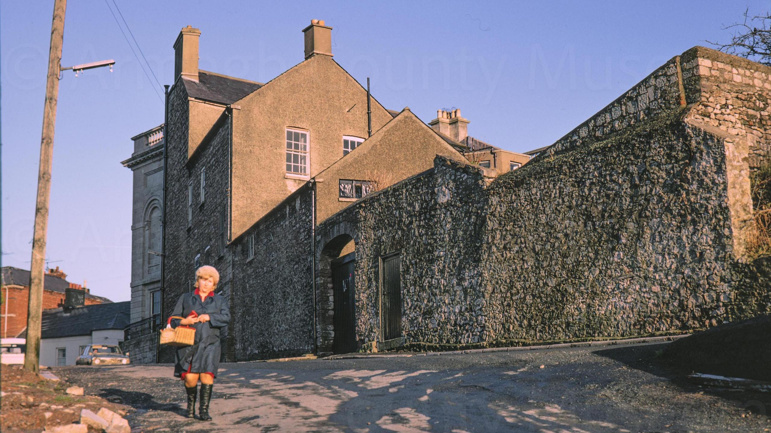 A digitalised photo from a film of an old stone building. A lady wearing a navy coat can be seen walking past. 
Looking north east from Callan street to Armagh Public Library. A woman in blue raincoat carrying a shopping basket walks towards the camera in the evening sunshine