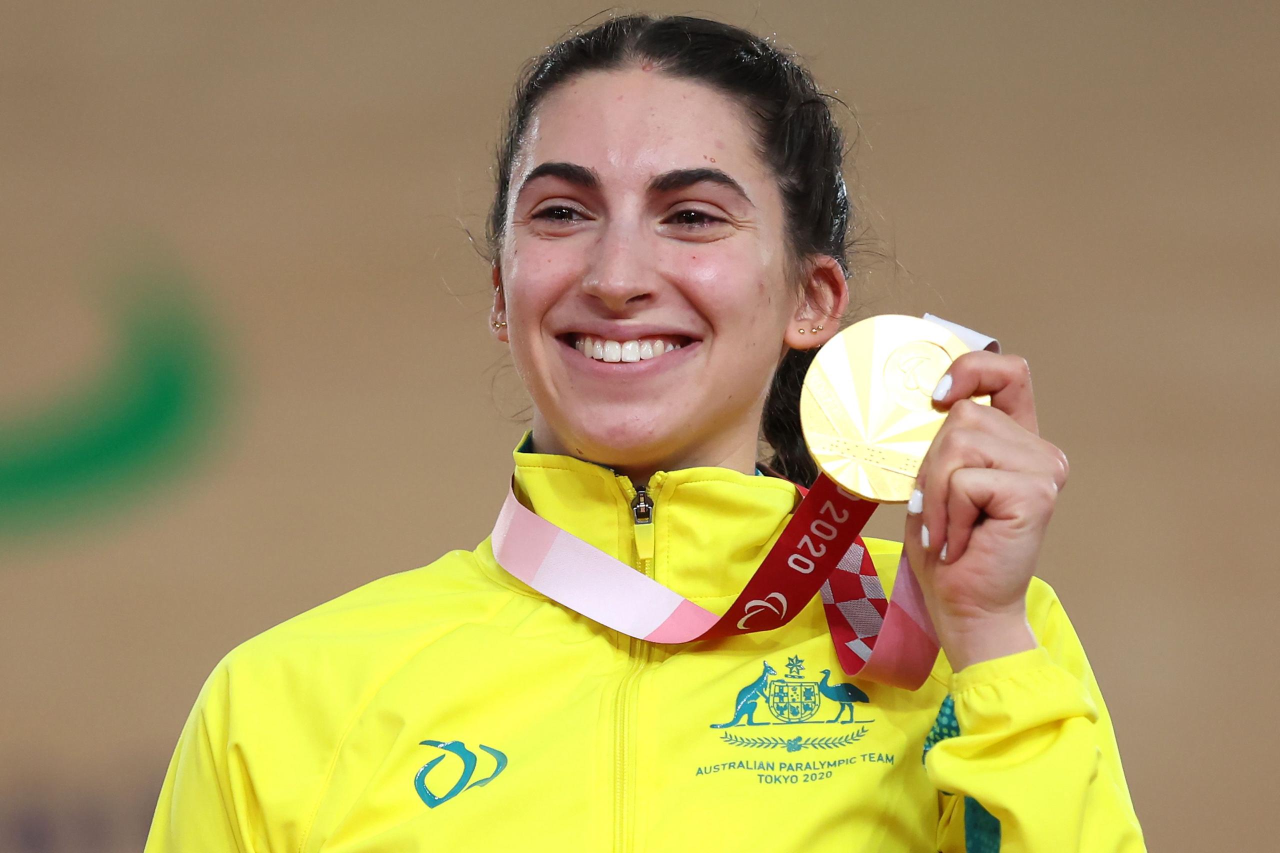 Paige Greco, e veshur me një xhaketë të verdhë të ndezur me logon e Ekipit Paralimpik Australian dhe tekstin "Tokio 2020", mban lart një medalje ari të bashkangjitur në një fjongo të kuqe dhe të bardhë. Medalja shfaqet dukshëm në plan të parë, dhe sfondi është pak i turbullt, duke sugjeruar një vend sportiv të mbyllur ose një vendosje në podium.