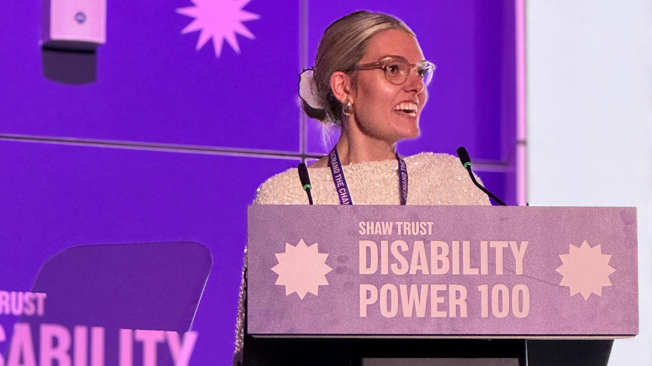 Celia Chartres-Aris speaking on the stage at the Disability Power 100 Awards show. She is wearing glasses.